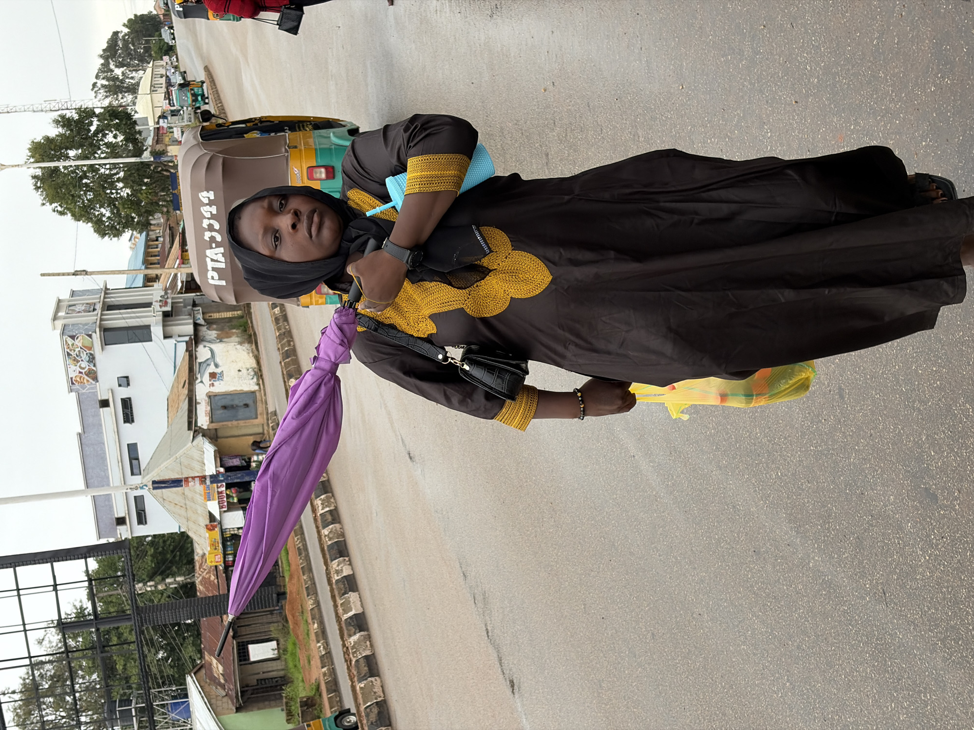 A person wearing a black hijab and a black and gold embroidered dress stands on a paved street. The person is holding a closed purple umbrella in their left hand and a yellow plastic bag in their right hand. A black purse is slung across their body. In the background, a three-wheeled vehicle, commonly known as an "Okada" or "Keke Napep," is visible. Buildings and trees line the street, suggesting an urban or semi-urban environment. The sky appears overcast, indicating a cloudy day. The overall scene suggests the person might be out shopping or running errands. The text "PTA-3344" is visible on the side of the three-wheeled vehicle. The location context is Jos, Nigeria.