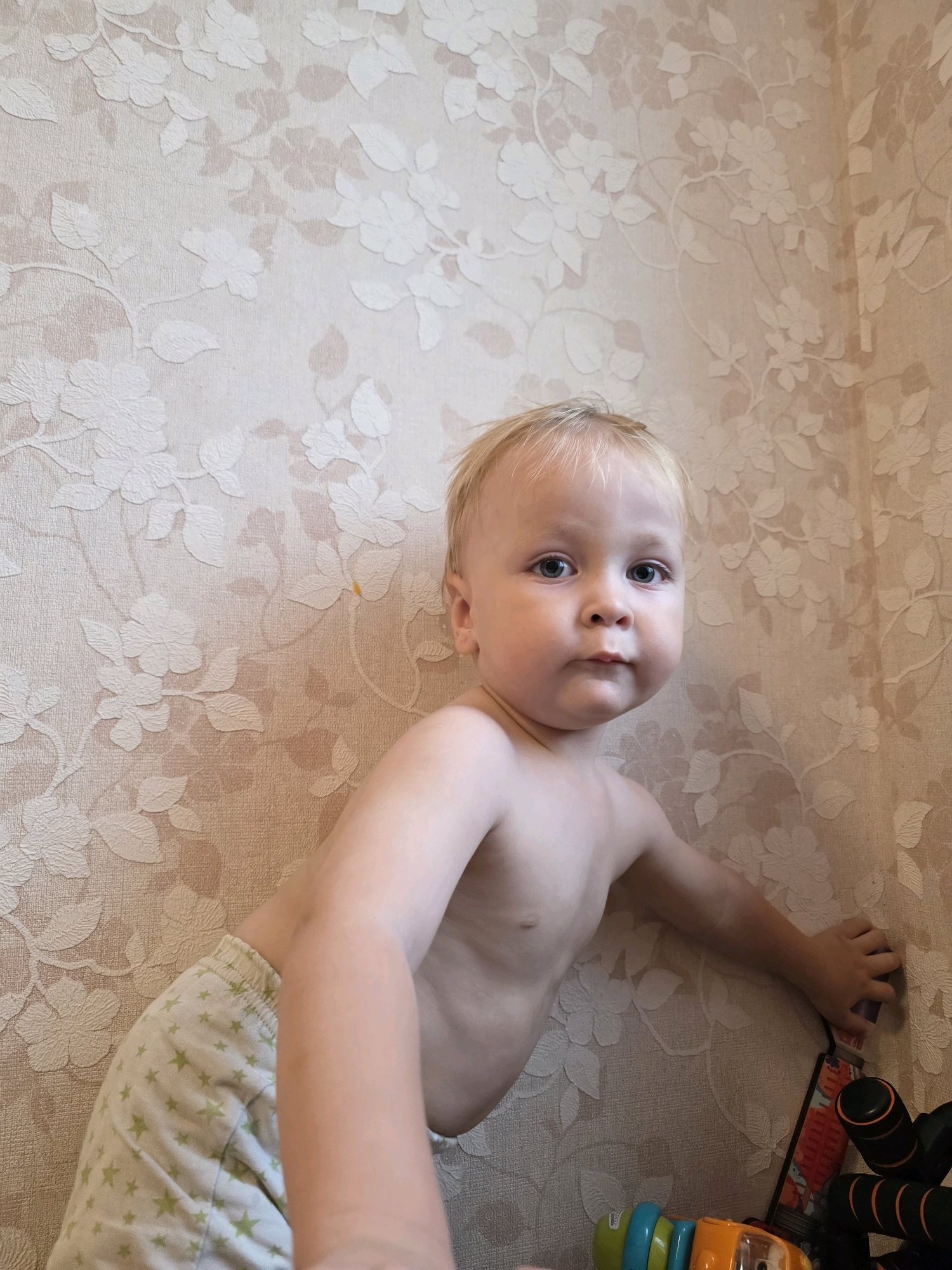 A young Caucasian boy with blonde hair and blue eyes is leaning against a beige floral wallpapered wall in Chaikovsky, Russia. He is shirtless, wearing light green shorts with a pattern of small, light green stars. His right hand is resting on the wall, and his left arm is extended forward. In the lower right corner of the image, several toys are visible, including a red and black toy that appears to be a toy vacuum cleaner or similar, and a colorful toy with blue, green, and yellow sections. The lighting suggests it is daytime. The boy's expression is neutral.