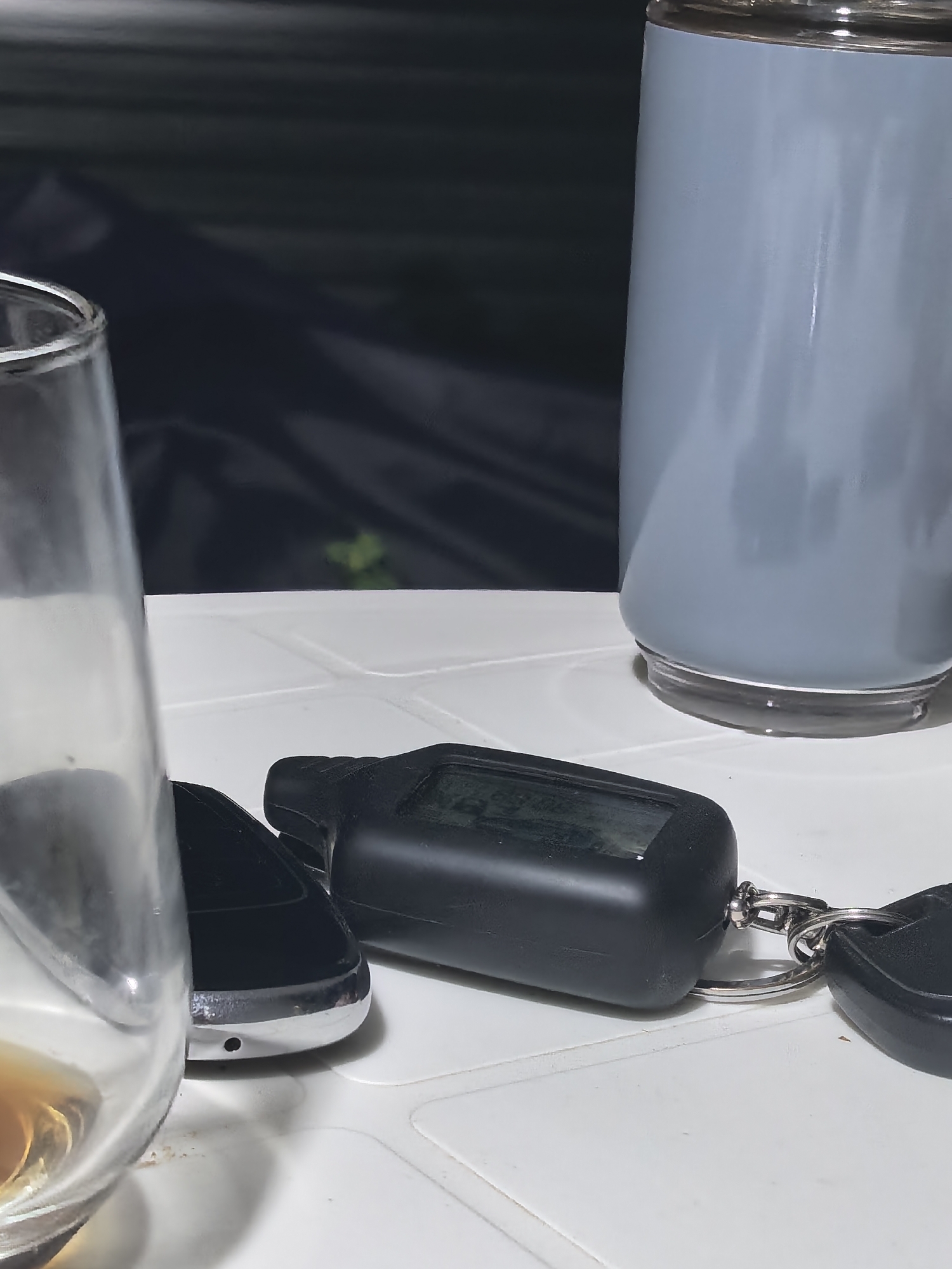 An outdoor table is laden with objects, suggesting a casual setting. In the foreground, a clear glass partially filled with a brownish liquid, possibly alcohol, is situated on the left. To its right, a black car remote key fob with a small screen and buttons lies on the white surface, attached to a keychain with a silver ring and another black object. Behind these, a tall, light blue tumbler stands upright. The background is dark and out of focus, hinting at foliage and possibly a dark tarp or fabric. The overall scene is still and appears to be captured in low light conditions. The location is identified as Beloyarskaya Zastava, Russia, though no specific urban landmarks are visible.