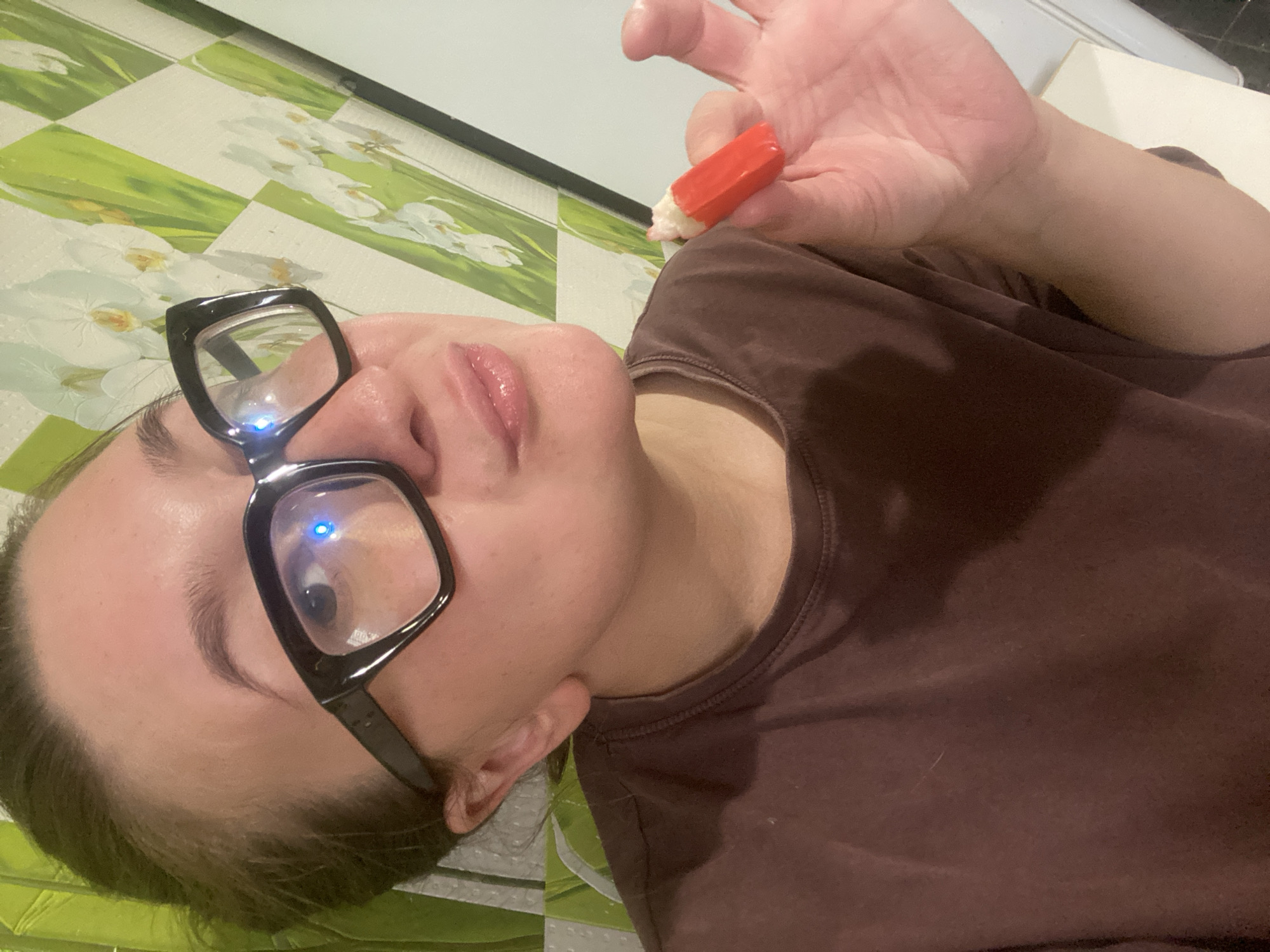 A person wearing black-framed glasses and a brown t-shirt is holding a red object with a white tip between their fingers. The person's face is partially visible, with their lips slightly parted. The background features a wall with a green and white checkered pattern and floral designs. The image appears to be a selfie taken indoors, possibly in a residential setting, and does not contain information indicating a specific location within the city of Chaikovsky, Russia.