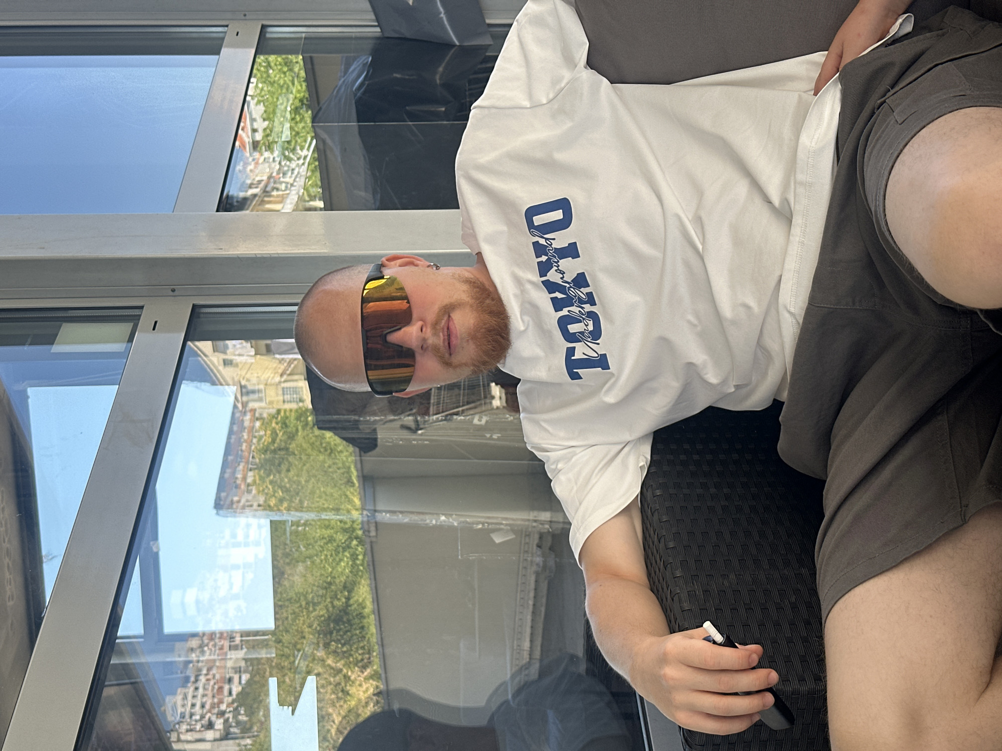 A bald man with a reddish-brown beard and mirrored sunglasses sits on a dark woven armchair. He is wearing a white t-shirt with blue text that reads "TOKYO Underground" and dark grey shorts. His left hand rests on the armrest, holding a white and black electronic cigarette device. The man is positioned in front of a large window with reflections of buildings and trees. The scene appears to be an outdoor seating area or a sunroom, possibly part of a cafe or residence in the city of Grad Rijeka, Croatia, given the architectural style of the buildings reflected in the window.
