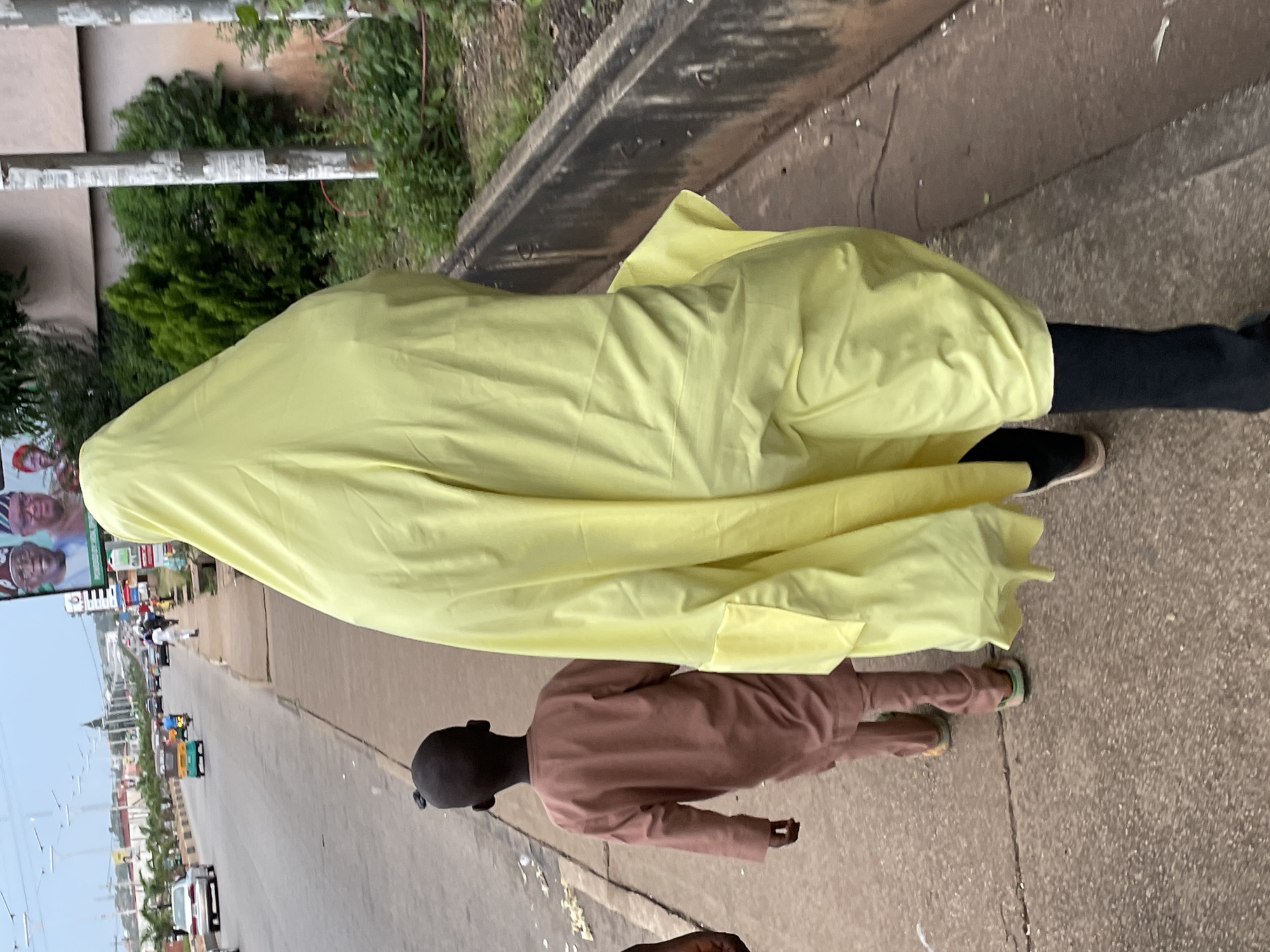 Two individuals are walking on a sidewalk adjacent to a busy road in Jos, Nigeria. The person in the foreground is wearing a flowing yellow garment that covers their entire body, including their head, obscuring their identity. To their left and slightly ahead, a young boy is walking, wearing a light brown, long-sleeved tunic and trousers. The road beside the sidewalk has several vehicles, including cars and a motorized tricycle, moving in the same direction. In the background, buildings and greenery are visible. A large billboard featuring images of people in traditional attire is also present. The sidewalk is paved and shows signs of wear, with cracks and scattered debris. The overall environment suggests an urban setting with moderate foot and vehicle traffic.