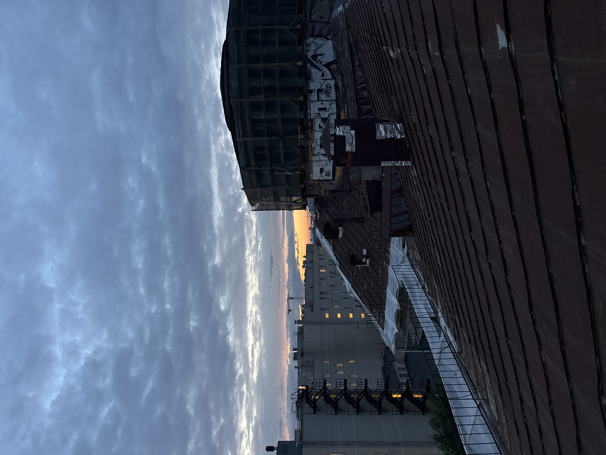 The image depicts a view of rooftops and buildings in Krasnoye Selo, Russia, during what appears to be either dawn or dusk. The sky is overcast with heavy clouds, but a sliver of orange and yellow light is visible near the horizon, indicating sunrise or sunset.
In the foreground, a rusted metal rooftop dominates the lower half of the frame. It is weathered and shows signs of age, with distinct lines formed by overlapping metal sheets. A dark chimney with metallic cladding is visible on this rooftop.
Behind the foreground rooftop, several other buildings and structures are present. To the left, a light-colored multi-story building features external metal fire escapes with stairs. Several windows on this building are illuminated with a warm, yellow light. Further in the background, other buildings stretch across the horizon, some with visible windows. A construction crane is also present in the distance, suggesting ongoing development or renovation in the area.
On the right side of the image, a large, circular building is partially obscured by scaffolding and netting. The facade of this building is covered with graffiti, predominantly in white and gray lettering. This structure appears to be either under construction or undergoing significant repair.
The overall scene is urban and somewhat industrial, with a mix of residential or office buildings and a prominent construction site. The lighting suggests a transitional time of day, contributing to a muted and somewhat dramatic atmosphere. No people are visible in the image.