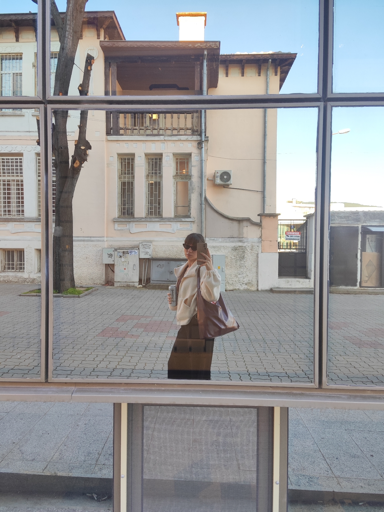 A reflection in a multi-paned window depicts a woman holding a phone, presumably taking a photograph. She is wearing sunglasses, a light-colored jacket, and a dark skirt or trousers. A large brown tote bag hangs from her shoulder. She also holds a drink container.
The reflection shows a street scene. The sidewalk is paved with interlocking stones. Behind the woman, in the reflection, is a building with multiple windows and a tree. There is an air conditioning unit mounted on the building's exterior. Part of the building features a balcony with a wooden railing. In the distance, there is a fenced area with a sign.
Based on the architectural style of the building in the reflection, the location may be in the city of Sliven, Bulgaria.