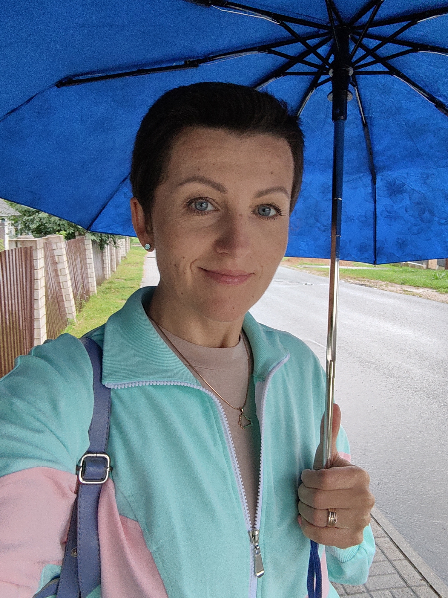 A medium-shot, front-facing selfie features a Caucasian woman holding a blue umbrella over her head. She has short, dark brown hair, blue eyes, and is smiling. She is wearing a light blue and pink jacket zipped up over a light brown t-shirt, with a silver necklace and a gold ring on her right hand. A blue purse strap is visible on her left shoulder. The background shows a paved road and a wooden fence with a grassy area and trees beyond. The scene appears to be outdoors in a residential area. This image was taken in Fanipal, Belarus.