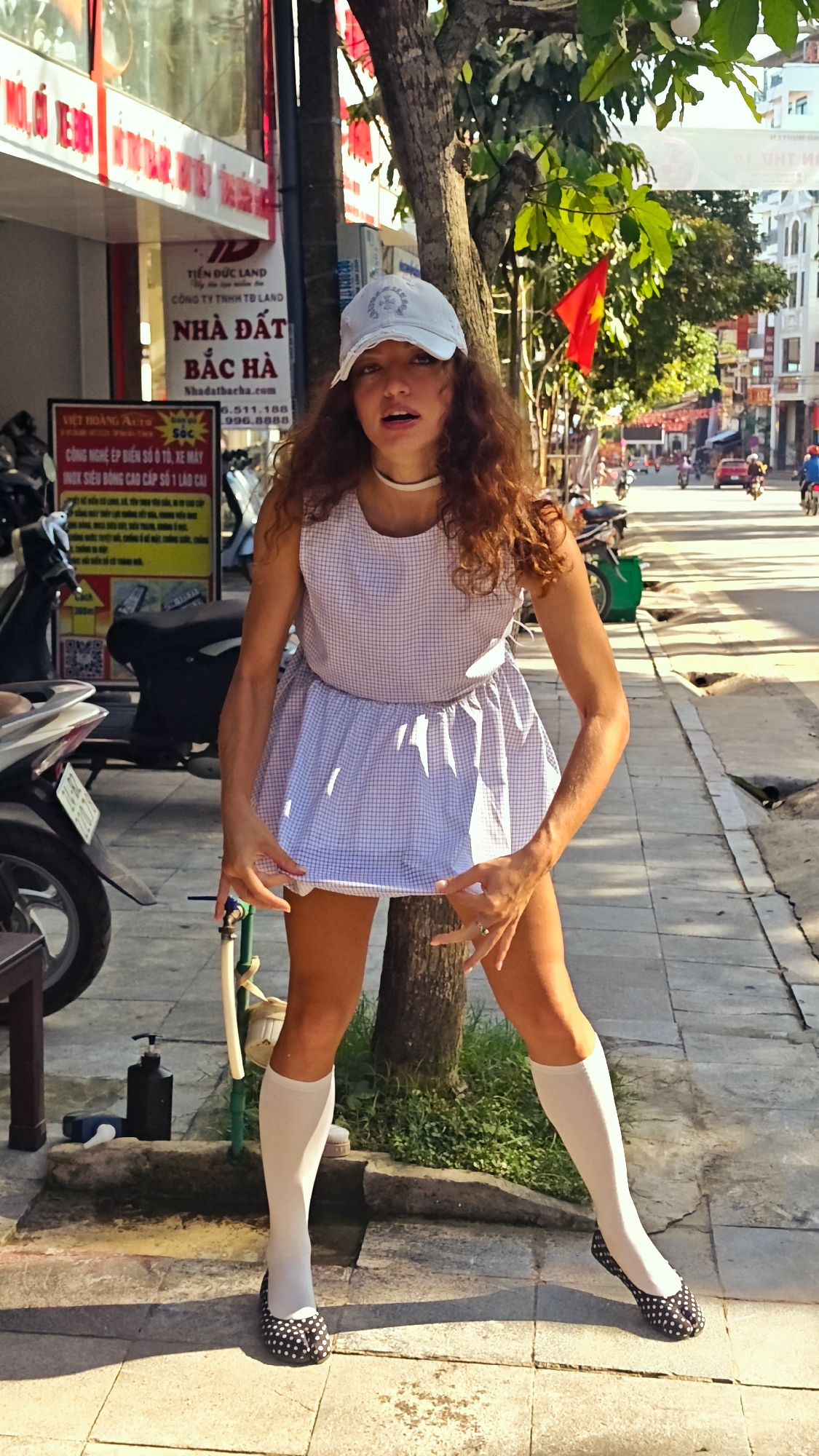 Here is a detailed description of the image:
**Subject:** A young, light-skinned person with curly reddish-brown hair is the central figure. They are wearing a light blue baseball cap, a white choker, a sleeveless, blue and white checkered dress, knee-high white socks, and black and white polka-dot flats. They are standing in front of a tree, holding up the hem of their dress with both hands, and looking directly at the camera.
**Location:** The scene appears to be a sidewalk in a city in Vietnam. Signs in Vietnamese are visible in the background, including one that reads "TIẾN ĐỨC LAND NHÀ ĐẤT BẮC HÀ" indicating that the location is in the city of Bắc Hà. There is also a sign that reads "VIỆT HOÀNG AUTO".
**Environment:** The sidewalk is made of light-colored stone. A motorcycle is parked nearby. A drainage area with some pipes and a bottle of soap are visible at the base of the tree. The background shows a street scene with buildings, trees, Vietnamese flags, cars, and other motorcycles moving in the distance.
**Action:** The person is holding up their dress hem, seemingly posing for the camera.
**Additional Details:** The photo is taken during the day with bright sunlight. The light casts shadows on the sidewalk and the person.