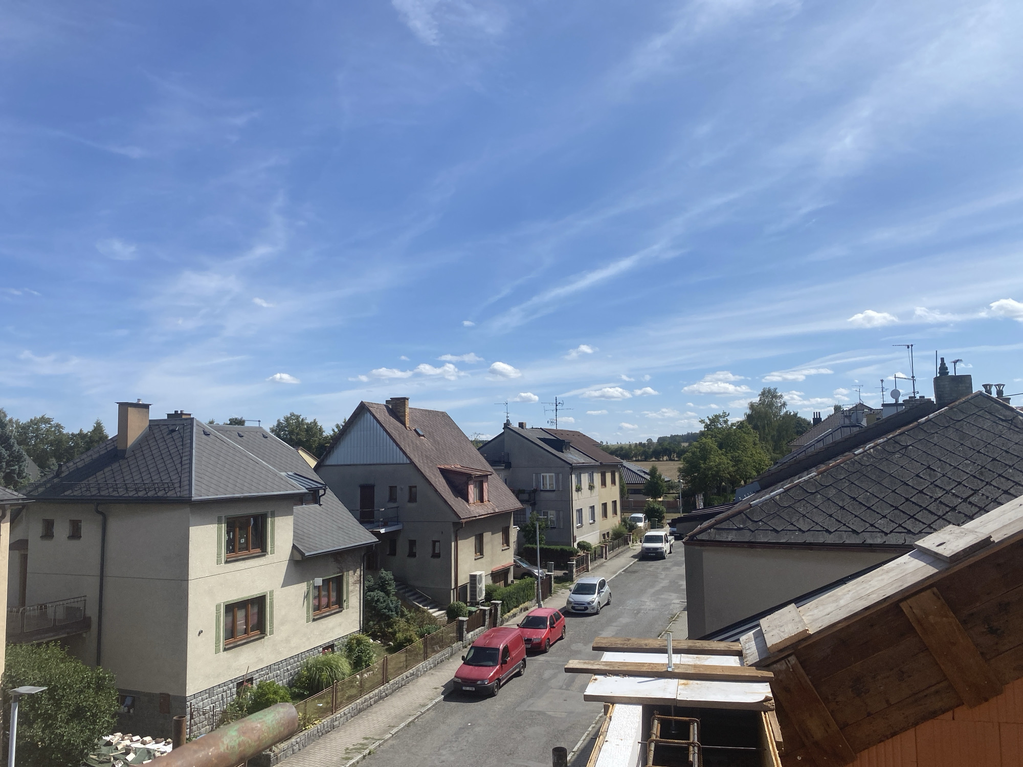 This is a high-angle, medium-shot, eye-level exterior photograph taken on a sunny day in Polička, Czechia. The scene depicts a residential street lined with houses and a clear blue sky with scattered cirrus clouds. In the foreground, a section of a rooftop under construction is visible, with wooden planks and building materials. The street itself is paved and curves slightly, with two red vans and two silver cars parked or driving on it. Houses of various architectural styles and colors are situated on either side of the street, some with green shutters on the windows and others with visible roof antennas. Trees and foliage are present on the sides of the road and in the background, suggesting a suburban or semi-rural setting within the town. The overall impression is a peaceful, everyday scene in a European town.