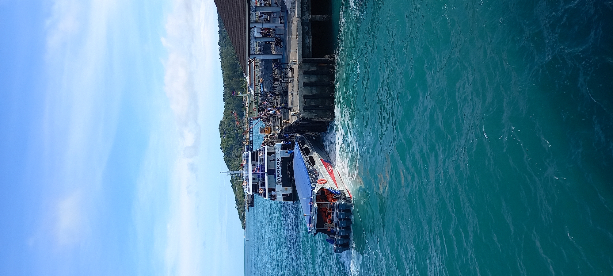 The image shows a ferry docked at a pier in ตำบลเกาะพะงัน, Thailand. The ferry, named "BANGKOK," is docked at a concrete pier with people standing on it. A green, wooded area is visible behind the pier. The water is turquoise and rippled. The sky is blue with some white clouds.