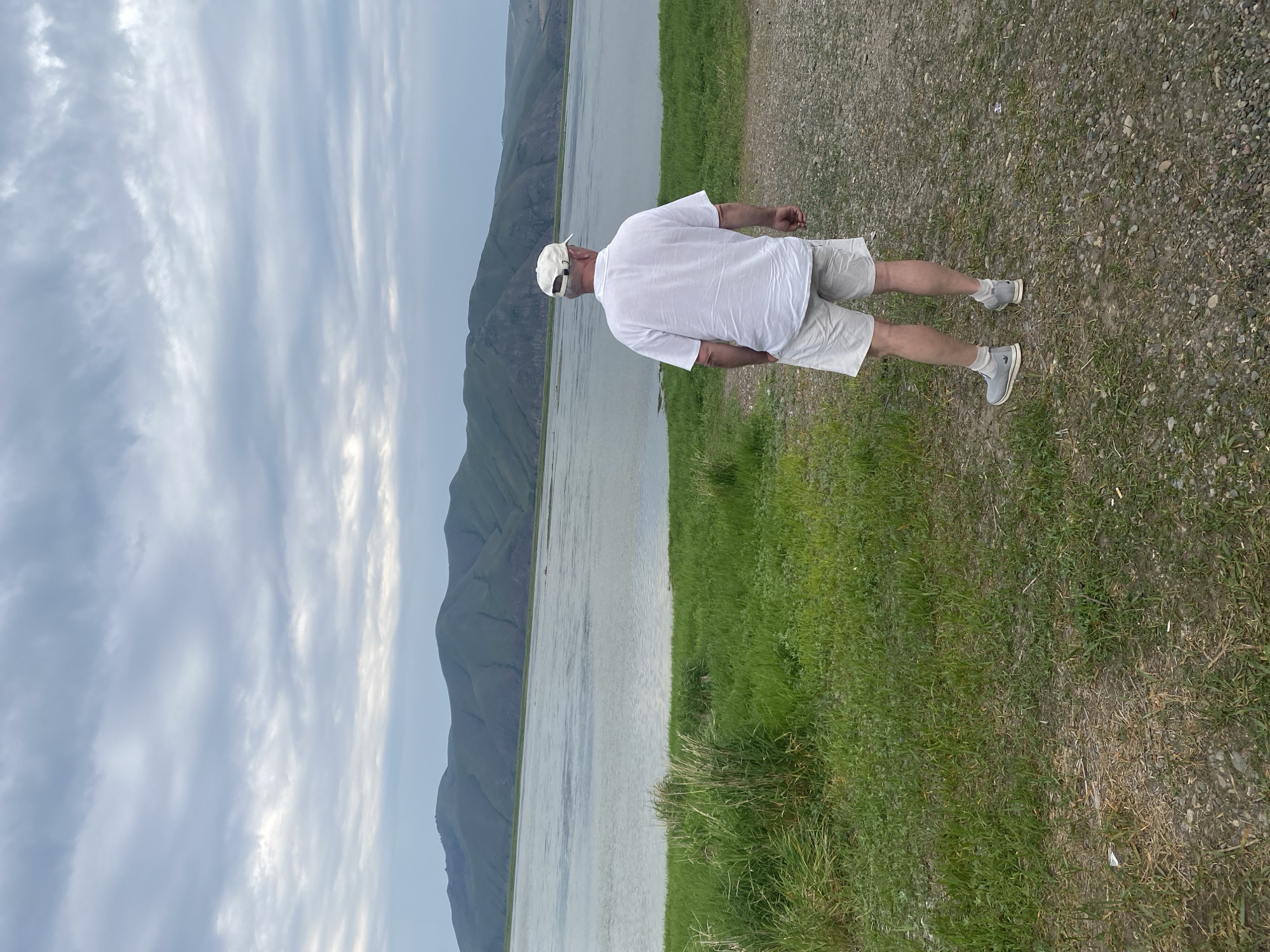 A man, viewed from behind, stands on a gravel path next to a grassy field, looking out over a large body of water towards a range of green, rolling hills under a cloudy sky. The man is wearing a white short-sleeved collared shirt, light-colored shorts, a white baseball cap, and grey slip-on shoes with white socks. The water is calm with some ripples, and the hills in the distance are covered in vegetation. The sky is overcast with soft, diffused light. This scene is likely located in or near the city of Ust-Abakan, Russia, given the landscape.