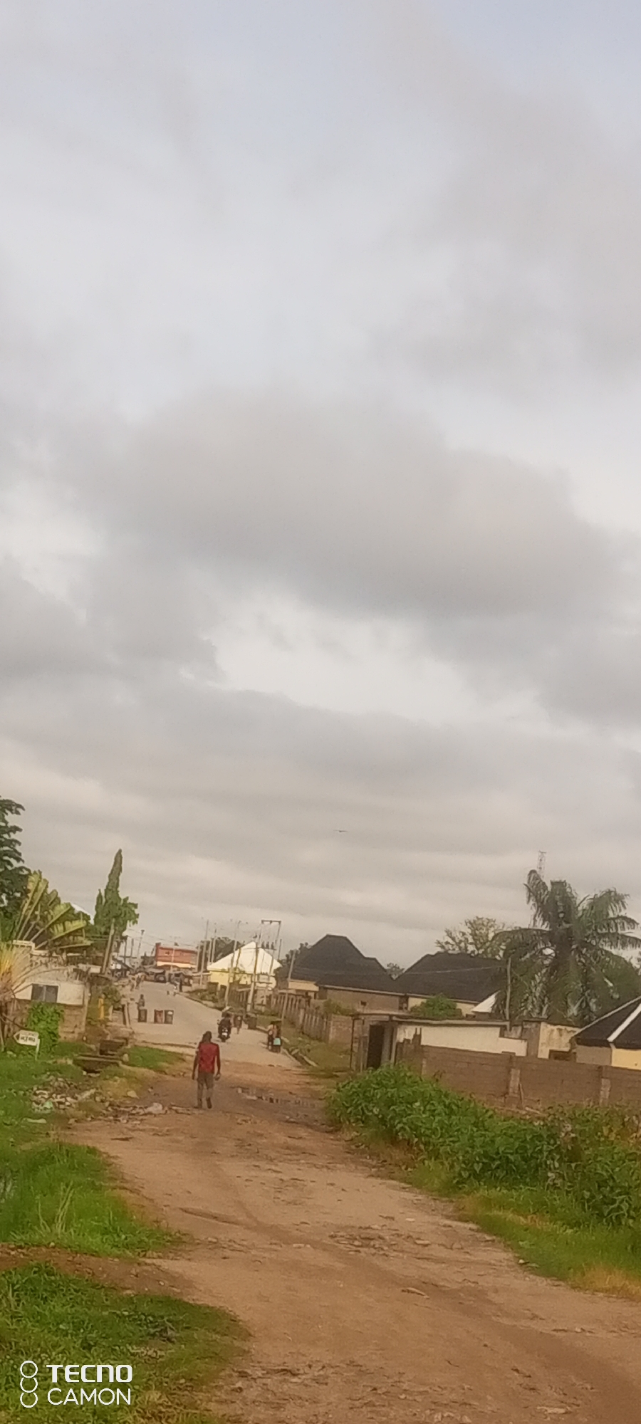 The image depicts a dirt road in a residential area, likely in Jalingo, Nigeria. A person wearing a red shirt walks away from the camera towards buildings in the distance. Several motorcycles are visible on the road, with some people riding them. The buildings appear to be single-story structures with tiled roofs, some with brick walls and others with lighter-colored facades. Vegetation, including grass and trees, is present along the sides of the road. The sky is overcast with clouds. The presence of the TECNO CAMON watermark suggests the photo was taken with a TECNO CAMON smartphone.