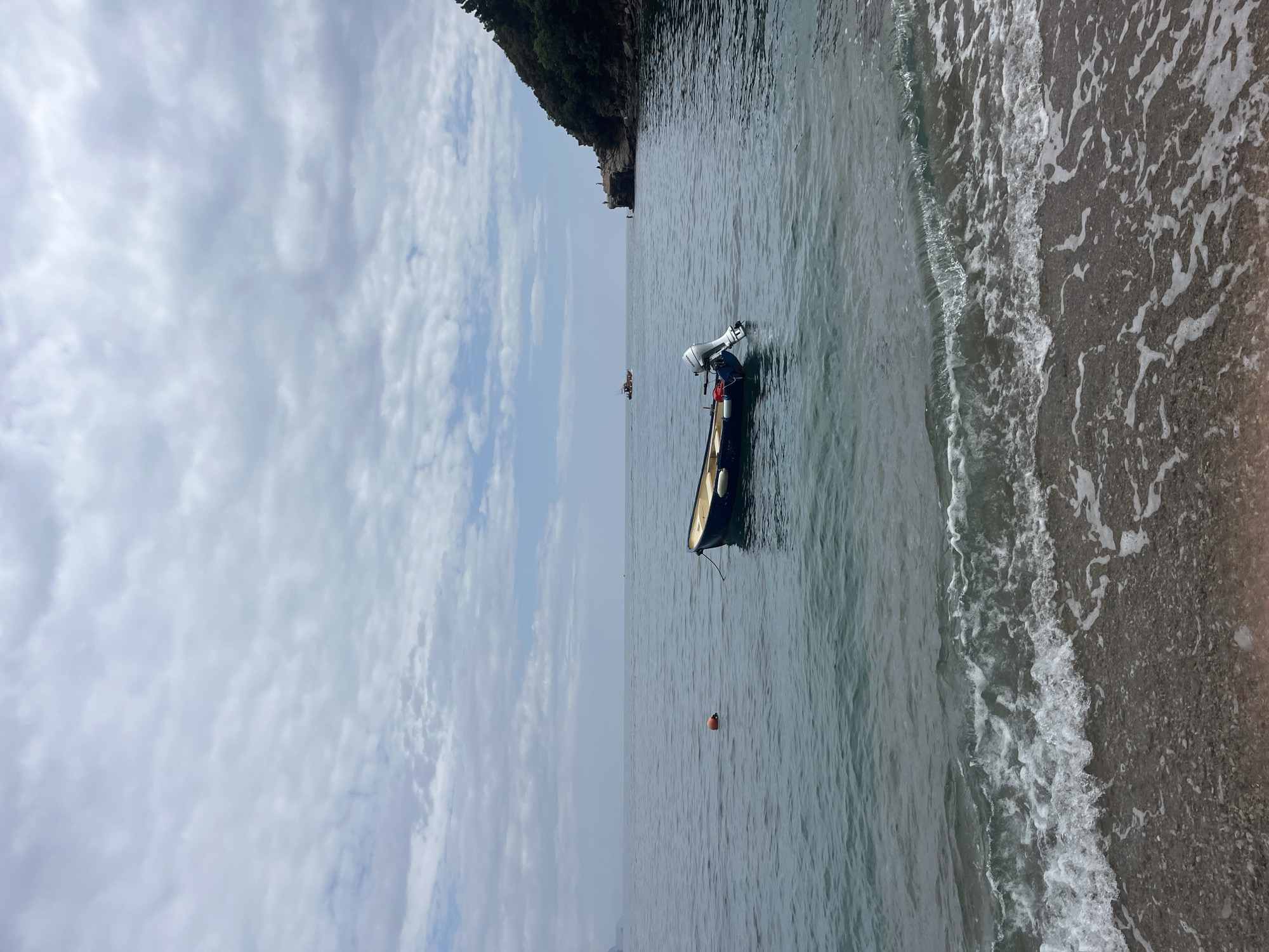 The image shows a view of the sea in Budva, Montenegro, with a boat anchored near the shore. The water is calm, and small waves are visible along the beach. There are clouds in the sky. A portion of a rocky cliff and forest is visible. The boat is a small rowboat equipped with a motor, suggesting it's used for fishing or short trips. A red buoy floats in the water near the boat. In the distance, a small, indistinct vessel can be observed on the horizon.
