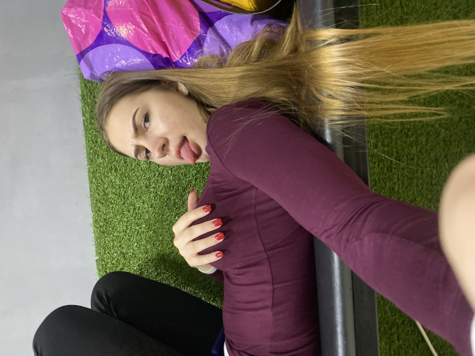 A young woman with long blonde hair lies on a surface covered in artificial grass. She is wearing a maroon long-sleeved shirt and black pants. Her right hand, with red nail polish, rests on her thigh. Her tongue is sticking out, and her facial expression suggests playfulness or a silly pose. Behind her, a large, colorful inflatable object, with pink and purple sections, is visible. The setting appears to be indoors, possibly a recreational space or studio, given the artificial turf and the presence of the inflatable. The lighting suggests it's daytime. The overall mood of the image is casual and lighthearted. There is no visible text in the image.