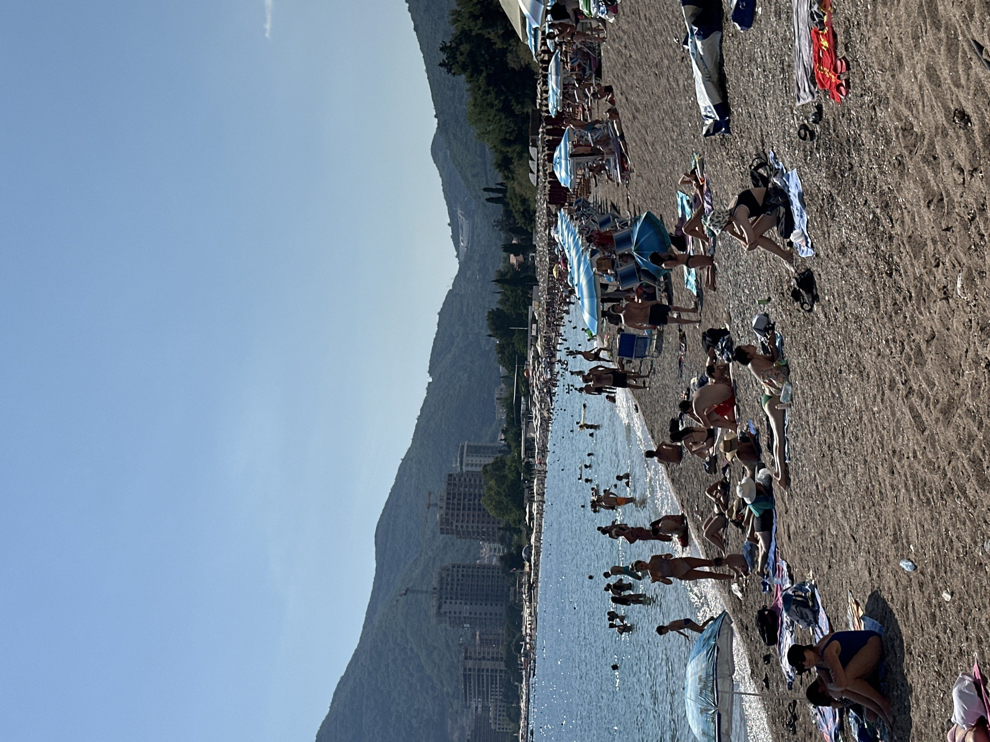 The image shows a crowded beach on a sunny day in Budva, Montenegro. The sea is calm and blue, with many people swimming and wading near the shore. The beach is made of pebbles and sand, with rows of blue and white striped umbrellas providing shade. Numerous people are sunbathing on towels and loungers, while others are walking along the shore. In the background, lush green mountains rise up, with modern buildings and construction sites visible on the slopes. The sky is a clear, bright blue.