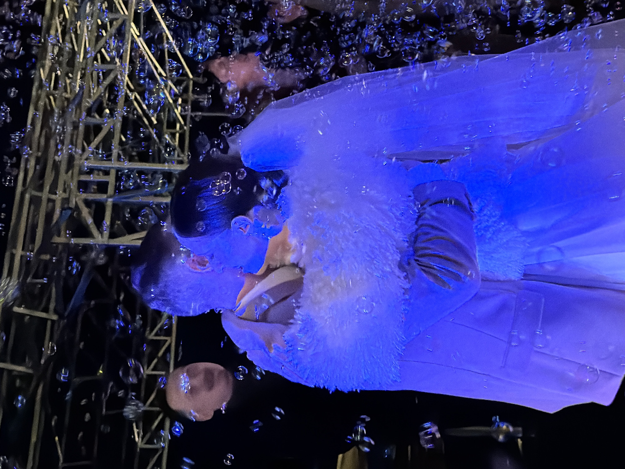 A couple in what appears to be wedding attire is shown kissing amidst a shower of bubbles. The scene is heavily bathed in blue light. Structural scaffolding is visible in the background on the left side. The exact location in Chaikovsky, Russia, is not discernible from the image.