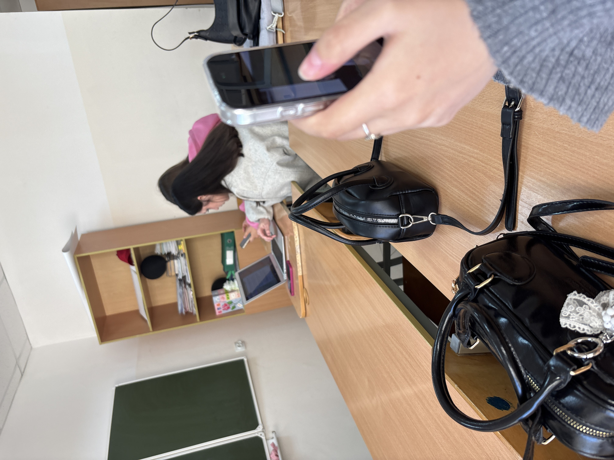 A student at the Chaikovsky Institute of Physical Culture in Chaikovsky, Russia, is photographing a black purse on a desk with their smartphone. Another student sits at a nearby desk with a laptop. A chalkboard and wall-mounted shelving are in the background.