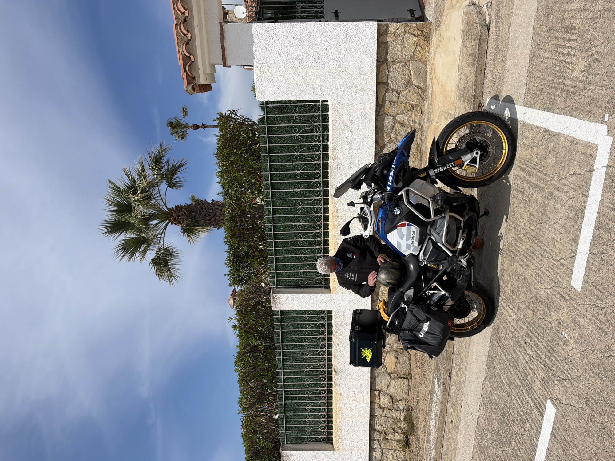 A man is stopped on a motorcycle in Mont-roig del Camp, Spain. The man, who appears older with short white hair, is wearing a dark jacket and sitting on a blue and white BMW motorcycle. He's holding a helmet in his hand. The motorcycle has black side cases attached. The scene is set on a paved street with white parking lines. Adjacent to the street is a low wall made of stone and white stucco, topped with a green metal fence partly obscured by shrubbery. A tall palm tree is visible beyond the fence, under a blue sky with scattered clouds. A building with a red-tiled roof is partially visible in the background.