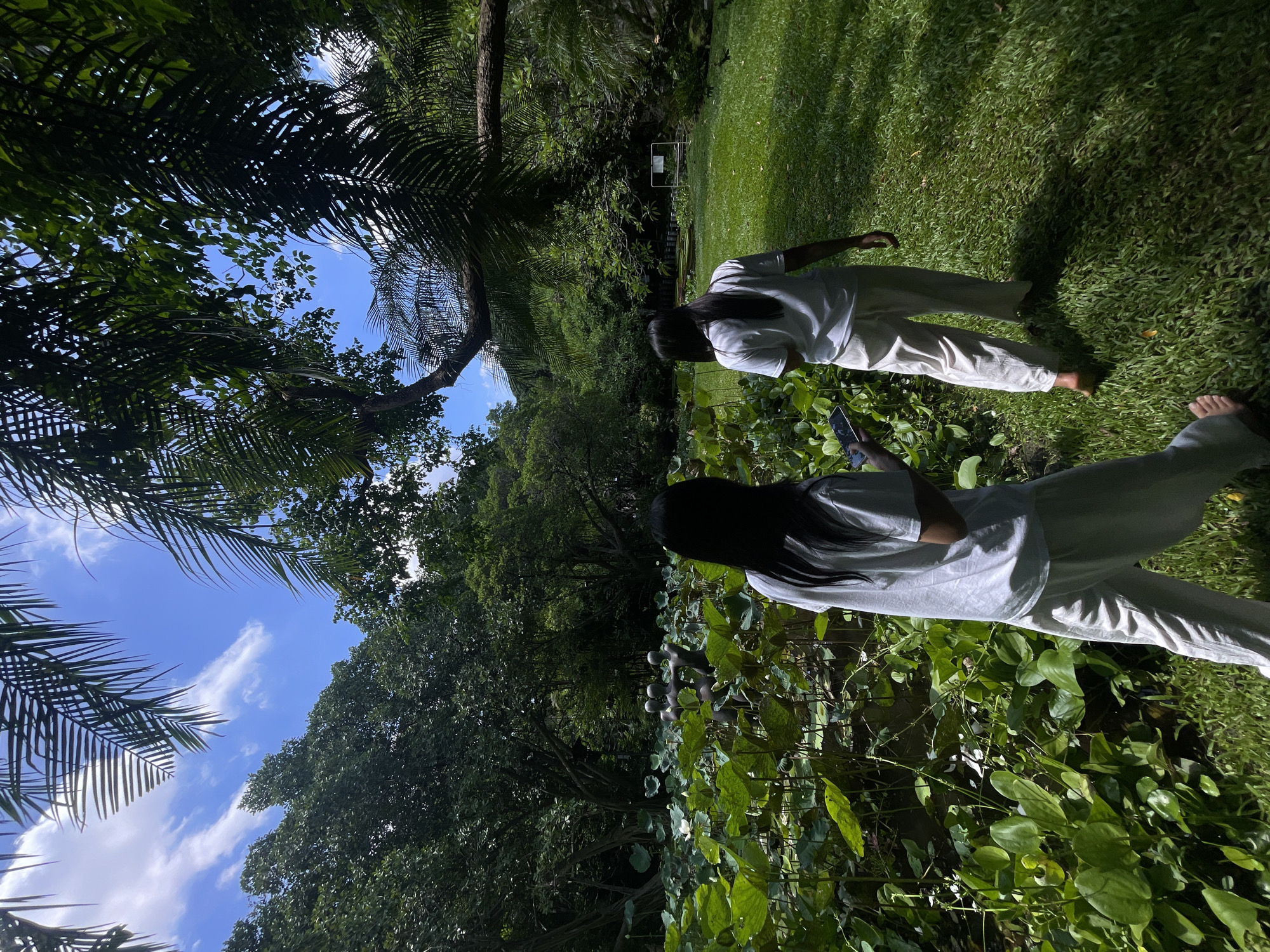 Two individuals, viewed from behind, walk across a lush green lawn in a garden setting. The person in the foreground is holding a smartphone. In the mid-ground, a pond is filled with large green lily pads, and a sculpture of three figures stands in the water. The background consists of dense foliage and trees. The sky above is blue with scattered white clouds. The scene appears to be the Bangkok Butterfly Garden and Insectarium in Chatuchak Park, Bangkok, Thailand, characterized by its tropical flora and outdoor exhibits.