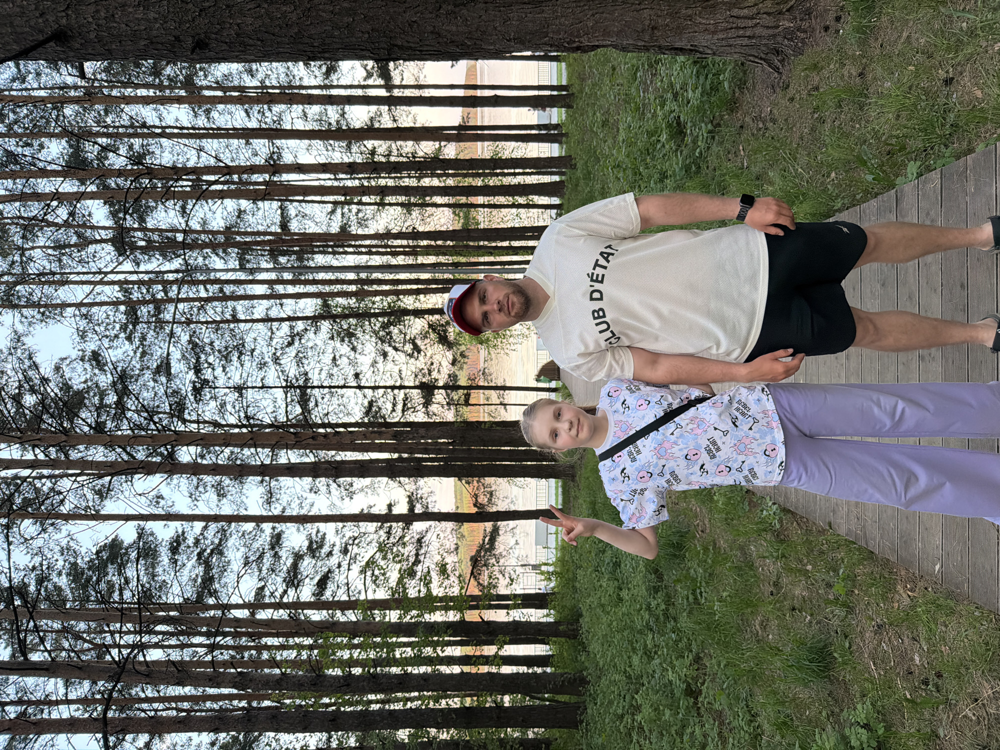 A medium outdoor shot shows a man and a girl standing on a wooden walkway through a grove of tall, slender trees, likely pine trees, with a glimpse of a body of water in the background.
The man, likely in his late 30s or early 40s, is wearing a white t-shirt with the words "CLUB D'ÉTAT" printed on it, black shorts, and a baseball cap. The girl, around 10-12 years old, is wearing a patterned t-shirt and lilac pants, and she is making a peace sign with her fingers.
The environment is a park or wooded area near water. The trees dominate the frame, creating a vertical pattern, while the wooden walkway provides a horizontal contrast. The presence of a body of water suggests this is near a river or lake. The grove seems well-maintained, given the established pathway. The location is Chaikovsky, Russia.