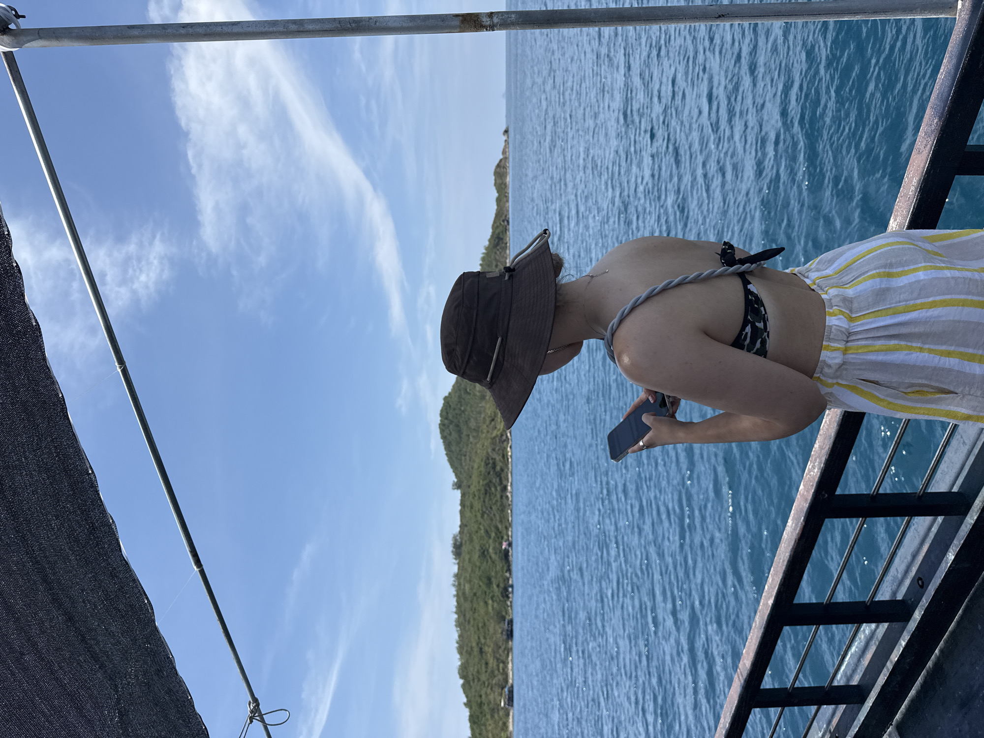 A person is standing on a boat with a brown hat and wearing a striped skirt. They are holding a black smartphone while looking towards a land mass in the distance. The boat has metal railings and a metal bar going across the top of the frame. The body of water, likely the sea, is blue. The sky is blue with wispy clouds. The land mass in the distance is covered with green trees. Given the location, the boat is likely in Nha Trang Ward, Vietnam.