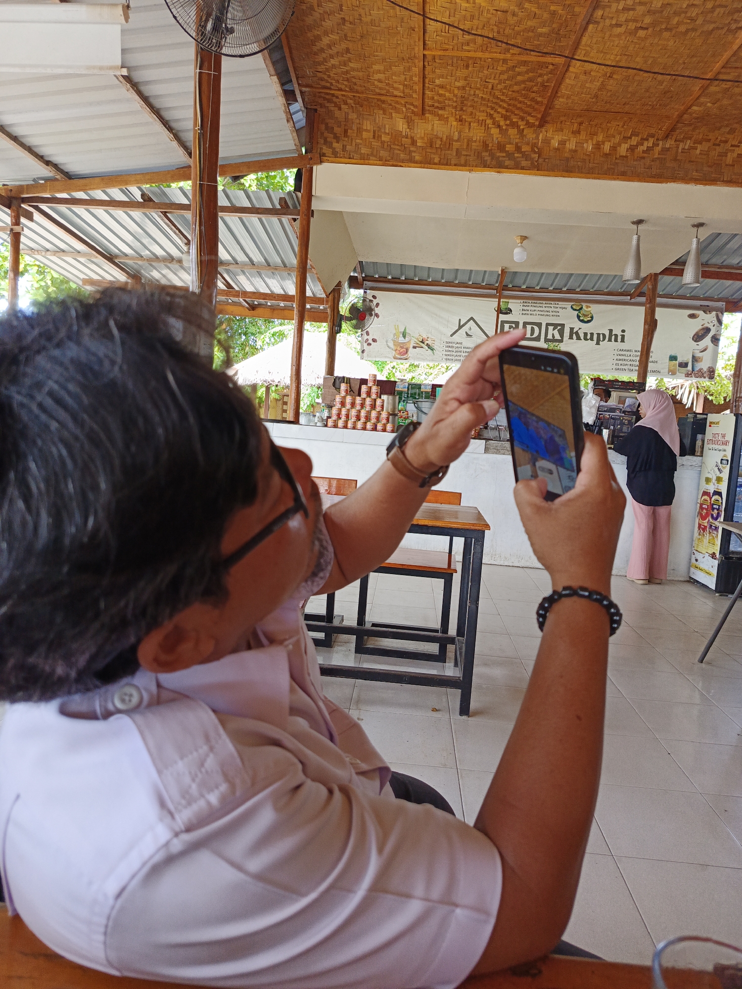 In Julikeude Dua, Indonesia, a person is taking a picture with their smartphone of a cafe's menu board. The scene is outdoors under a thatched roof and corrugated metal covering, with wooden beams supporting the structure. Tropical foliage is visible in the background, suggesting a warm climate. Sunlight streams in, illuminating the tiled floor and the cafe's interior.
The person in the foreground, viewed from behind, wears glasses and a light pink collared shirt. They are holding a smartphone in their right hand, capturing an image of the menu. The menu board, featuring text in Indonesian, lists various drinks such as "SEREH JAHE" (lemongrass ginger), "CARAMEL MA," and "ES KOPI." The cafe's name, "FDK Kuphi," is prominently displayed.
In the background, a woman wearing a pink hijab and a black top stands behind a counter, seemingly serving customers or preparing drinks. Stacks of canned goods and bottles of beverages are visible on shelves behind her, along with signage for "TASTE THE EXTRAORDINARY." The overall atmosphere appears to be casual and relaxed, typical of a daytime cafe setting. The image captures a moment of everyday life, showcasing the cafe's offerings and the patron's activity.