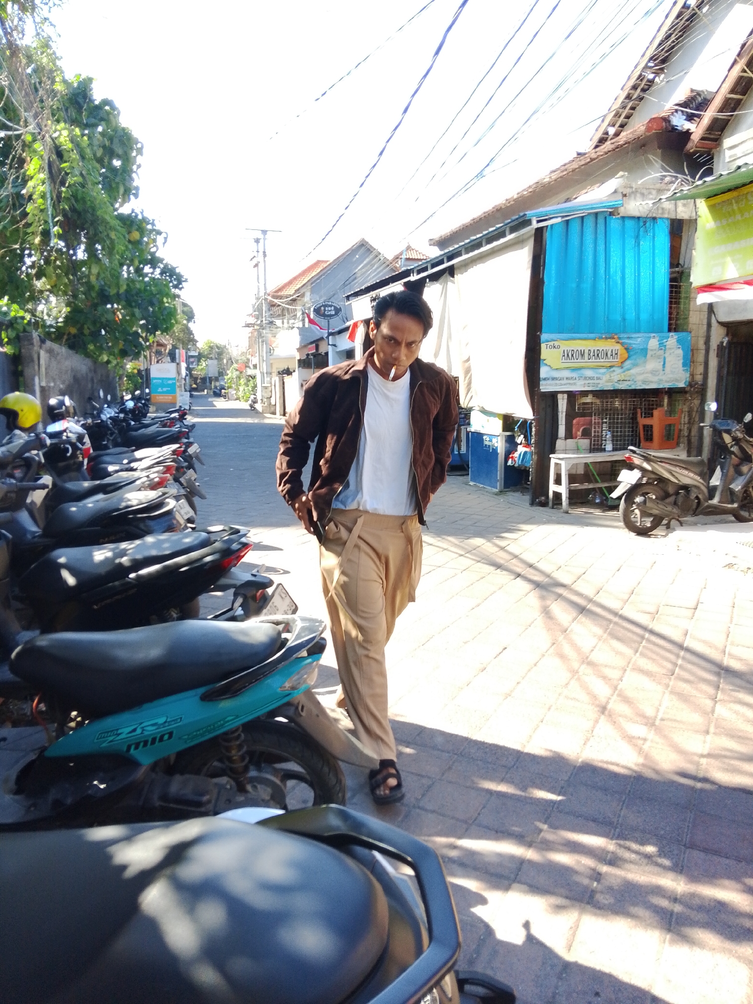 A man walks down a sidewalk in Kuta, Indonesia, holding a cigarette. He is wearing a brown jacket, a white t-shirt, and tan pants. The sidewalk is paved with interlocking bricks and lined with parked motorcycles. A building with a blue awning is visible in the background, along with a sign that reads "AKROM BAROKAH." The scene is illuminated by bright sunlight, casting shadows on the ground.