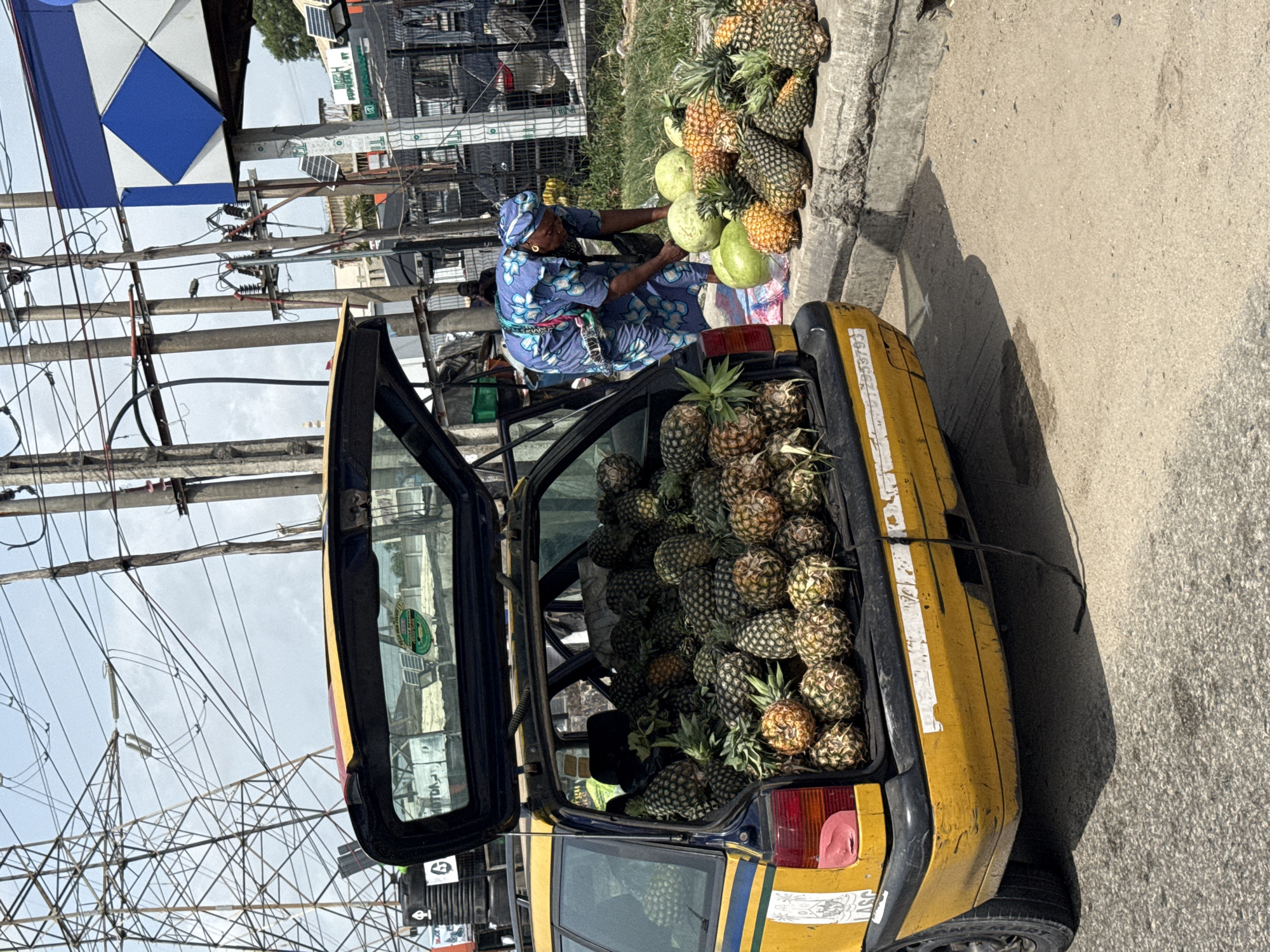 In Lekki, Nigeria, a woman in a blue patterned dress and headwrap tends to a roadside fruit stand. The stand includes a pile of pineapples and several watermelons. An open hatchback of a yellow taxi, filled to capacity with pineapples, is parked next to the stand. Overhead, there is a network of electrical wires and a blue and white sign. The ground is paved.