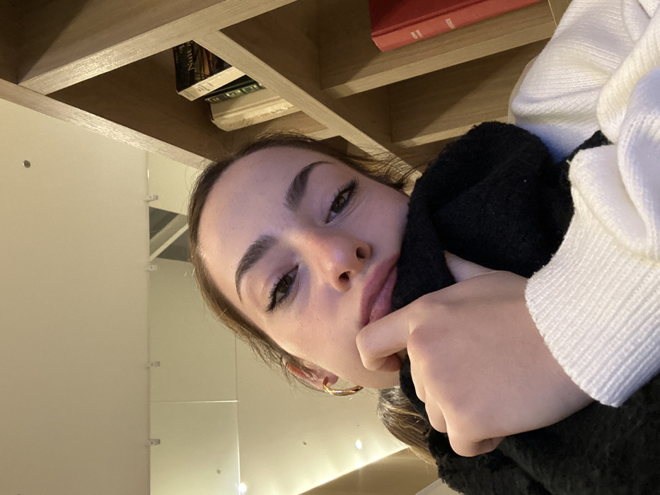 A young woman poses, head tilted, with a contemplative expression. Her hand is near her mouth. She is wearing a white sweater and a black scarf. The background includes a wooden shelf with books and a bright white wall with mirrored panels. The lighting is soft and warm. The image is taken from a low angle.