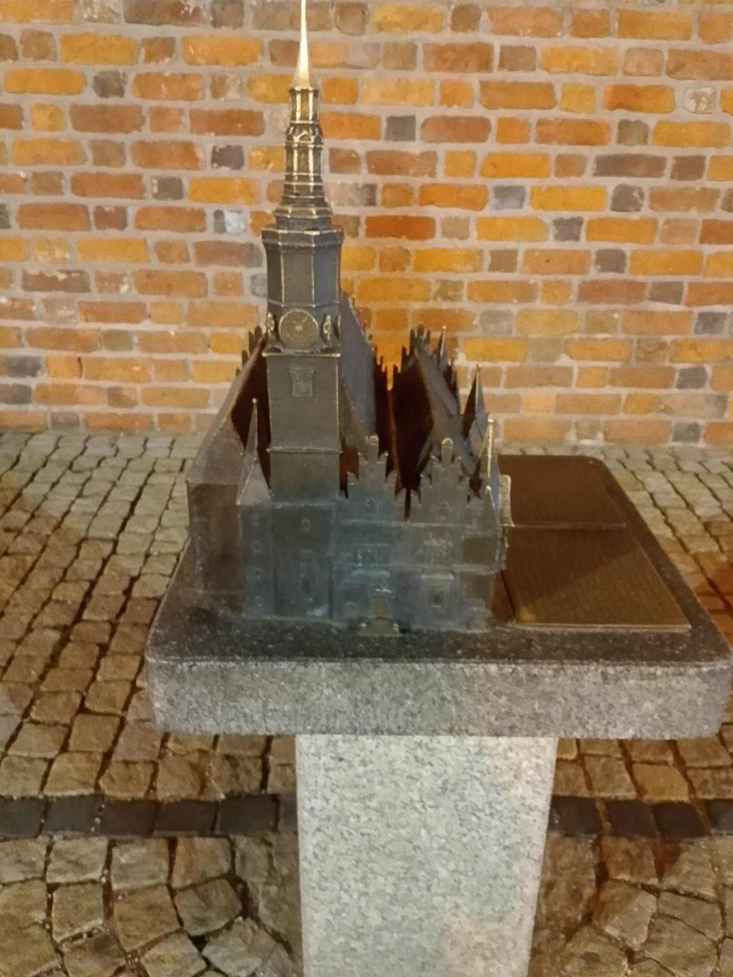 The image shows a miniature replica of the Market Hall (Hala Targowa) in Wrocław, Poland. The replica is made of dark metal and stands on a stone pedestal, which is placed on a cobblestone ground. The background is a brick wall. The Market Hall is a historic building in Wrocław, built in 1908. It is a two-story building with a distinctive Art Nouveau style. The replica shows the characteristic features of the building, including its tall tower and intricate facade. The cobblestone ground and brick wall suggest that the replica is located outdoors, possibly in a public square or park in Wrocław.