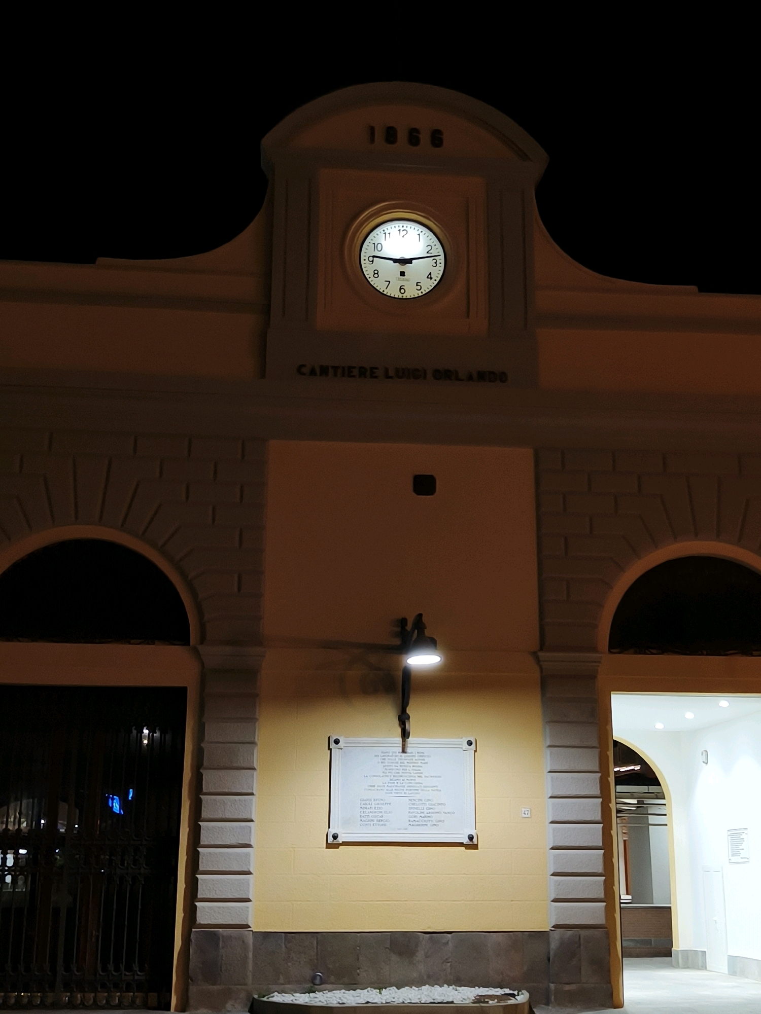 The image shows the entrance to the Cantiere Luigi Orlando in Livorno, Italy, at night. A large, ornate archway forms the main entrance, with two smaller arched openings on either side. Above the entrance is a clock tower with the year "1866" inscribed at the top. The clock face is illuminated and shows the time as approximately 2:15. The building is made of yellow brick and stone, with decorative brickwork around the arches. A single lamp post with a bright white light is mounted on the wall below the clock. To the left of the entrance, a dark metal gate blocks access to a courtyard. To the right, the entrance leads into a brightly lit interior with a vaulted ceiling. A plaque with text is mounted on the yellow wall below the lamp post. There are no people visible in the image.