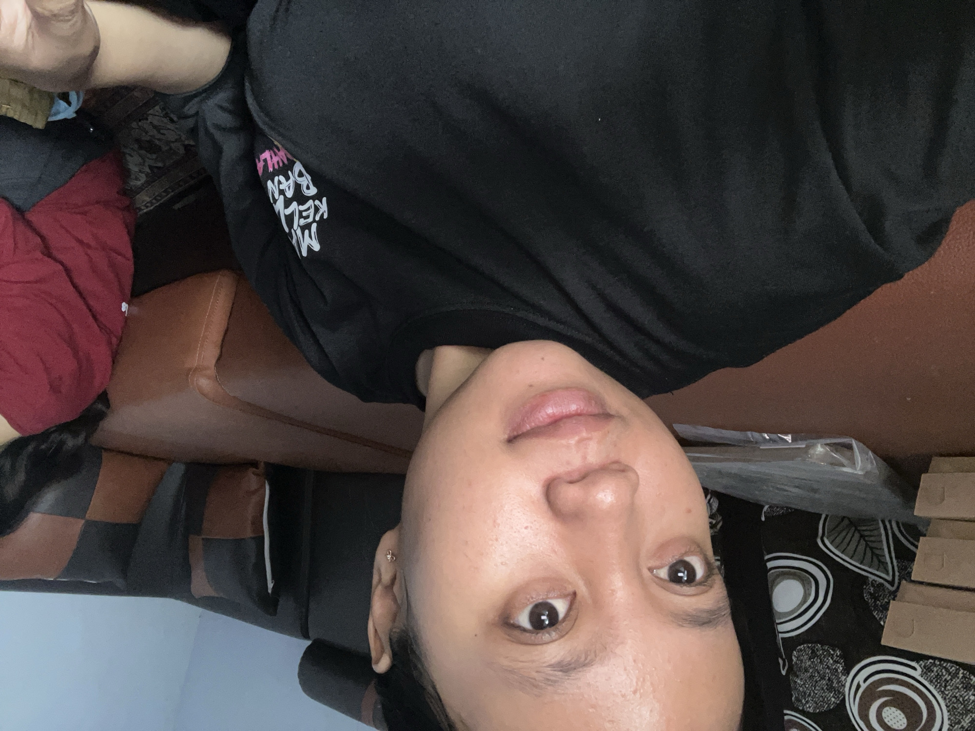 A young woman, appearing to be of Southeast Asian descent, is captured in a close-up, front-facing selfie. She has medium-toned skin, dark hair pulled back, and is wearing a black t-shirt with white and pink text that is partially visible. Her expression is neutral. The background consists of a brown leather sofa with dark grey and brown checkered pillows. To the left of the sofa, there is a stack of brown cardboard boxes and a plastic-wrapped item. The overall setting suggests an indoor environment, possibly a living room. The lighting appears to be from an artificial source, casting a warm glow on the subject.