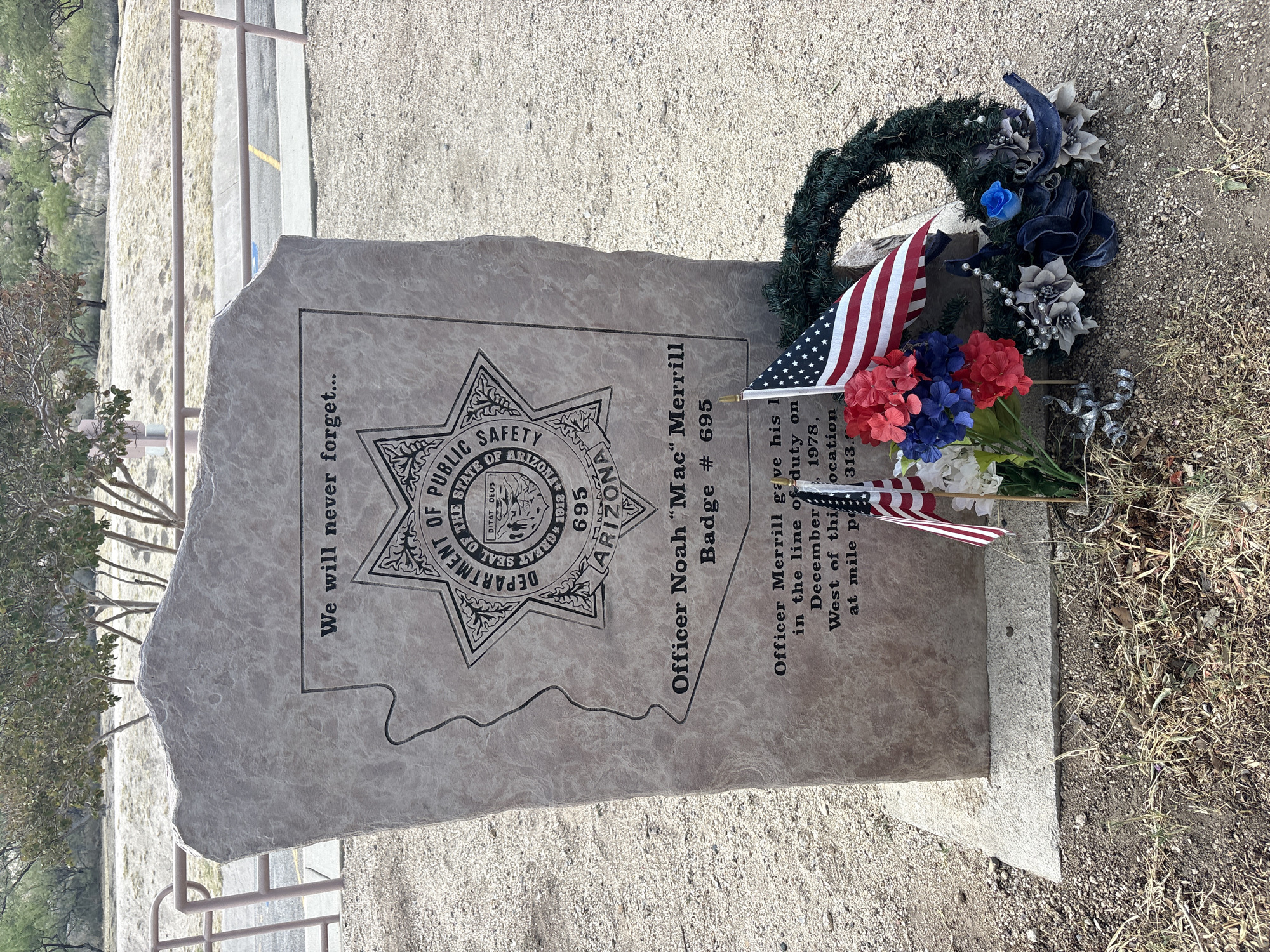 In the city of Dragoon, United States, a memorial stone stands in an outdoor setting. The stone is engraved with the outline of Arizona and the Department of Public Safety badge, along with the inscription "We will never forget..." and "Officer Noah 'Mac' Merrill Badge # 695". Below this, text reads, "Officer Merrill gave his l in the line of duty on December 1978, West of this location at mile po 313.6". At the base of the stone, a wreath and artificial flowers are placed. Two small American flags are positioned in front of the wreath and flowers. The ground surrounding the memorial is covered with gravel and dry vegetation, with a metal railing and trees visible in the background.