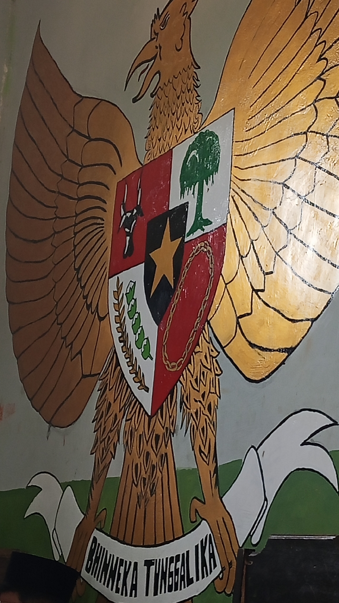 The image displays a mural of the coat of arms of Indonesia, the Garuda Pancasila. The Garuda, a mythical bird, is depicted with golden wings spread wide. In the center of the mural, a shield is emblazoned with symbols representing the five principles of Pancasila: a star (belief in one God), a chain (humanitarianism), a banyan tree (unity), a bull's head (democracy), and stalks of rice and cotton (social justice). Below the shield, a white banner displays the national motto "Bhinneka Tunggal Ika" (Unity in Diversity) in black lettering. The mural is painted on a wall in Suka Harapan, Indonesia, as indicated by the presence of a black peci (cap) worn by a person partially visible at the bottom left of the frame. The background of the mural is a light gray wall with a green stripe along the bottom. The overall image is a close-up, slightly angled view of the mural.