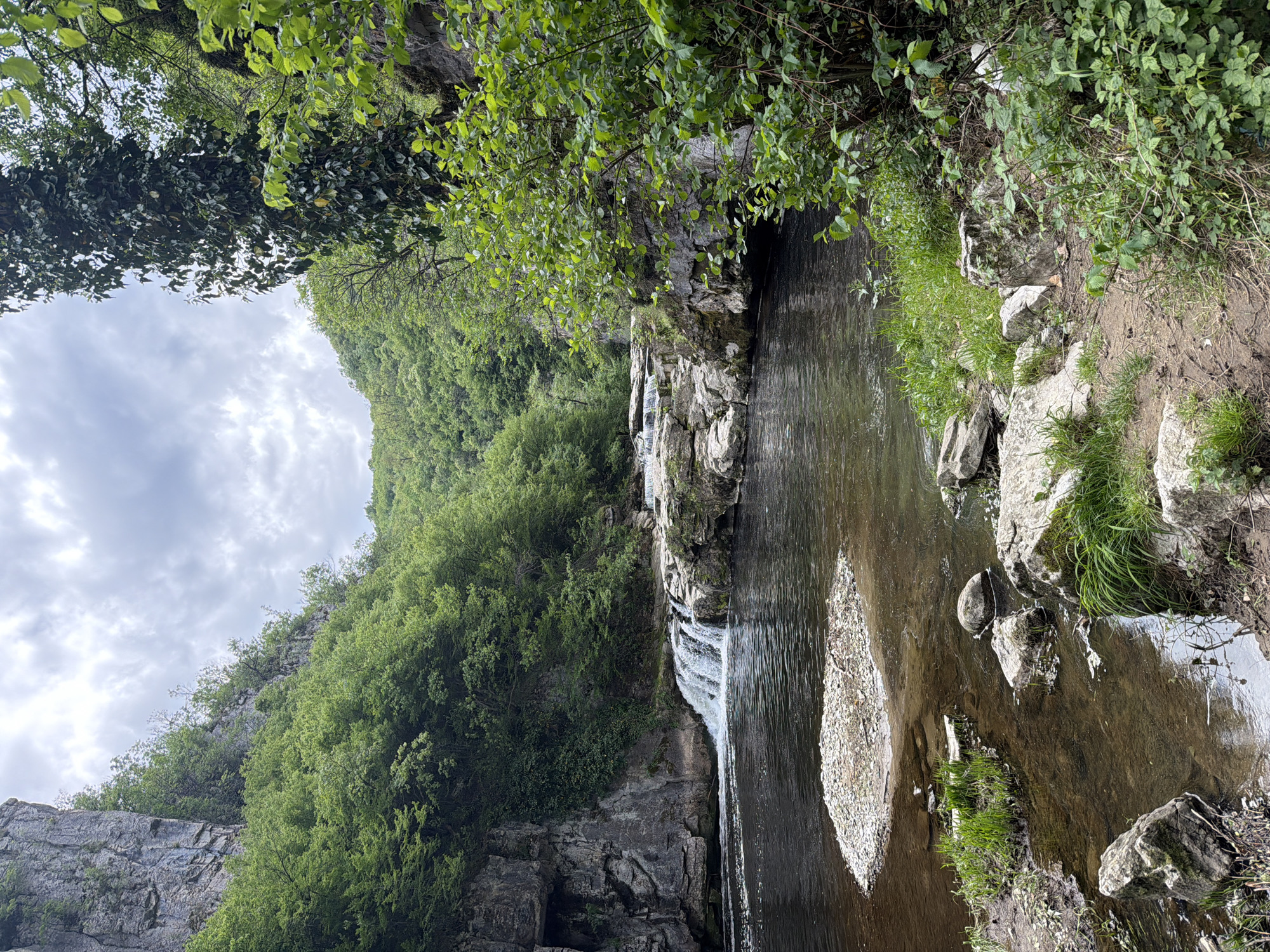 The image depicts a waterfall and a small, shallow river in a natural environment. The location is identified as Gesha, Bulgaria. The water flows over a series of rocky ledges, creating a cascade effect. The riverbed is visible, with a small island of pebbles and rocks. Lush green vegetation, including trees and grass, covers the surrounding hills and cliffs. The sky is overcast with cloudy conditions. There are no people visible in the image.