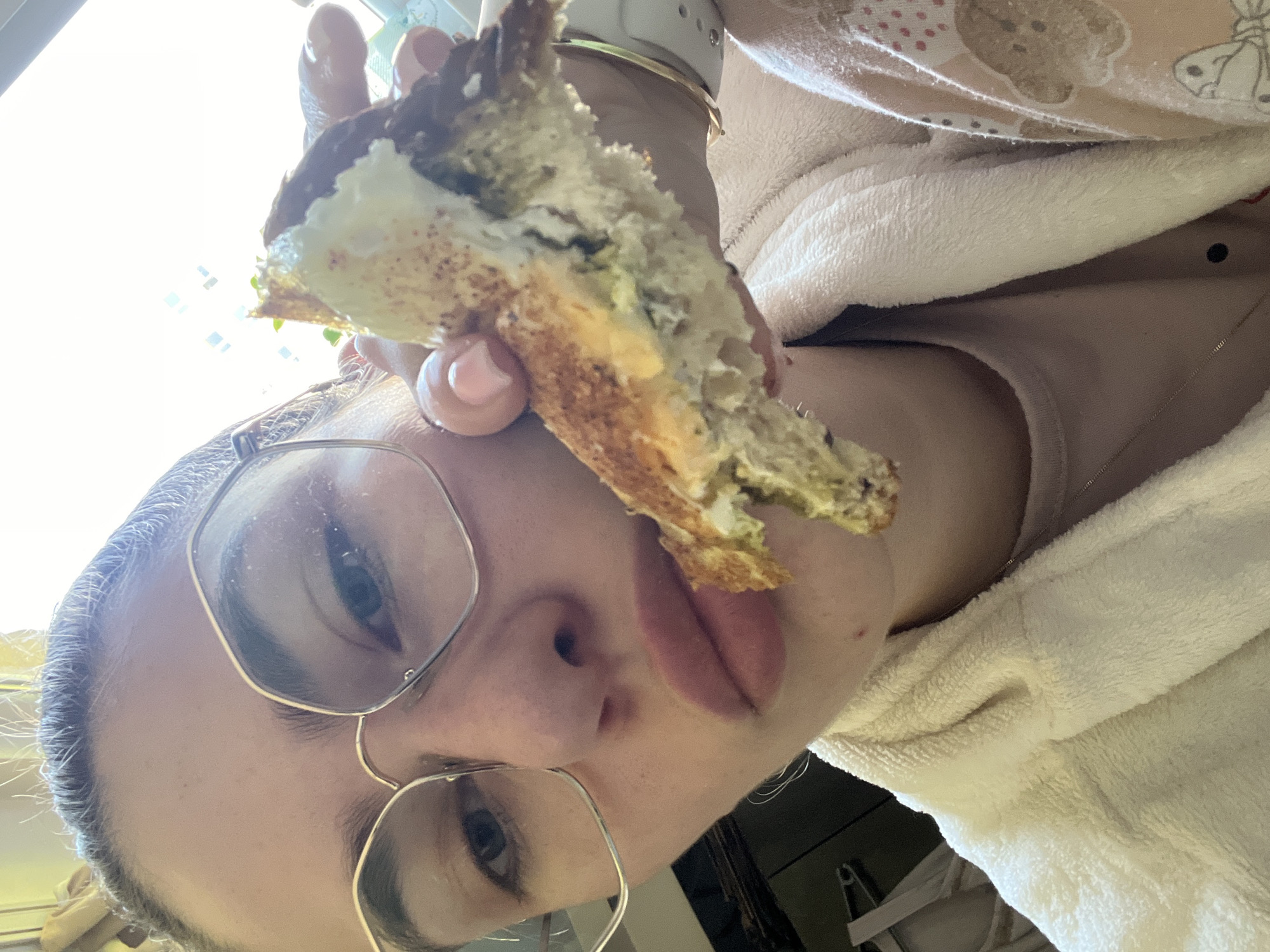 A low-angle close-up shows a person holding a half-eaten sandwich. The person has fair skin, glasses with gold frames, and pink lipstick. They are wearing a light-colored robe over a light-colored top, and a gold bracelet and smartwatch are visible on their wrist. The sandwich has a browned crust and a light-colored filling with green flecks. The background is out of focus, but a light source appears to be coming from a window. The location and city are Unknown, Unknown.