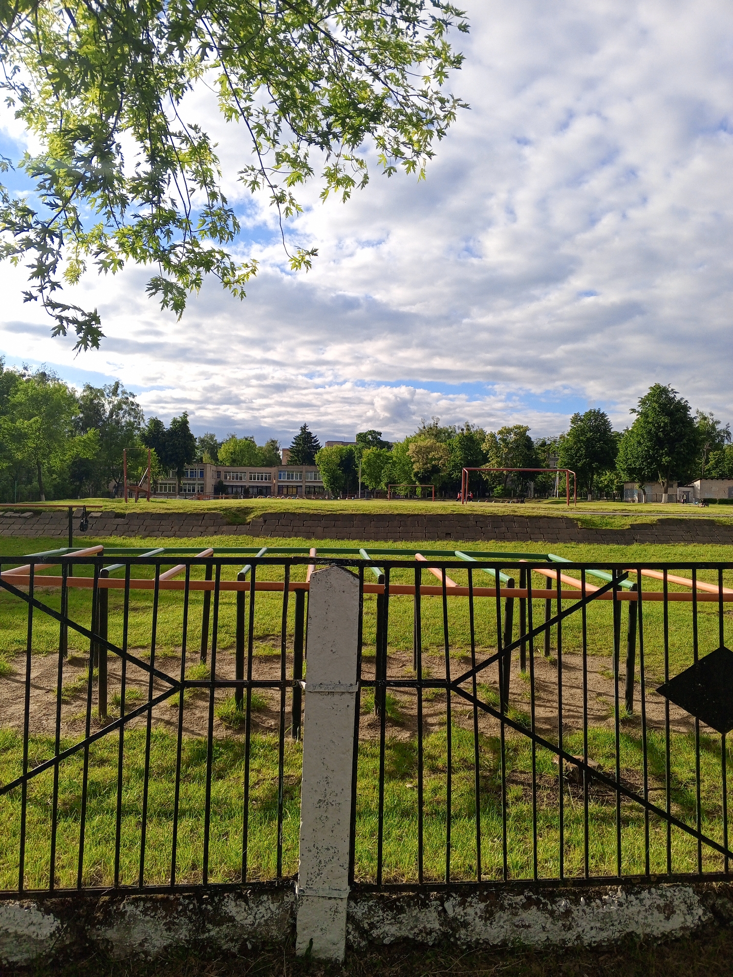 The image displays an outdoor scene, likely a schoolyard or public recreational ground, with the exact location in the city of Unknown, Unknown. In the immediate foreground, a black metal fence with vertical bars and a diamond-shaped accent on the right is present, supported by a worn concrete post, partially painted white. Below the fence, a weathered concrete base is visible.
Behind the fence, a patch of green grass interspersed with bare soil features several low-lying metal bars, painted in alternating green and orange, arranged as an obstacle or climbing structure. Further back, a retaining wall, constructed from stacked concrete or stone blocks, spans the width of the midground. Beyond this wall lies a expansive grassy field, possibly a sports pitch, where two red-painted goalposts, resembling soccer goals, are clearly visible; one on the right and another partially obscured on the left. A tall, reddish-brown metal structure, possibly a swing set or climbing frame, stands to the left on the field.
In the background, a large, multi-story building with numerous windows and a light-colored facade is situated behind the field and a line of various deciduous trees with dense green foliage. This building appears to be an institutional structure, such as a school or community center. Distant figures, possibly one or two people, are barely discernible near the rightmost goalpost. The sky above is partly cloudy, displaying patches of blue sky interspersed with white cumulus clouds. The lighting indicates daytime conditions, likely late morning or afternoon.