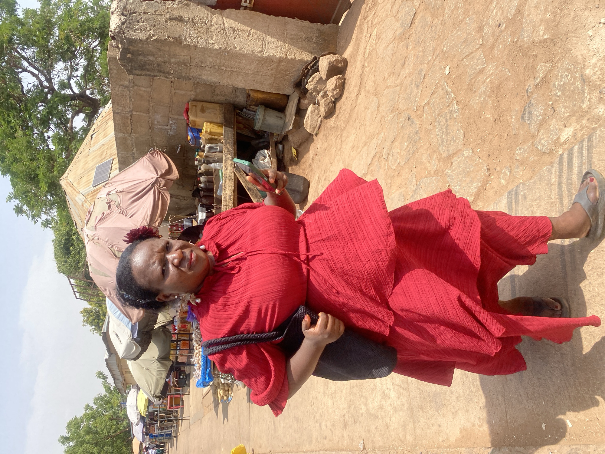 An outdoor scene in Rido, Nigeria, depicts an adult female walking on an unpaved path with embedded stones. She wears a red textured dress with ruffled sleeves, grey flip-flop sandals, and carries a black woven bag on her left shoulder. Her dark hair is styled up, adorned with a red floral accessory, and she wears gold-colored dangle earrings with white pearl-like drops. She holds a green-backed smartphone in her right hand and looks directly at the camera with a neutral expression.
To her left, in the background, a row of market stalls with various colored umbrellas (white, olive green, and light brown) shelters displays of goods, including piles of small, light-colored produce items. To her right, a weathered concrete block structure with a partially collapsed upper section stands. Behind this structure, a building with a corrugated metal roof features a small solar panel. Underneath a wooden shelf attached to the concrete structure, numerous plastic bottles (brown, clear), jerrycans (yellow, blue), and a grey bucket are displayed. The ground is mostly dirt with scattered rocks. Green trees are visible in the background against a light blue sky.
