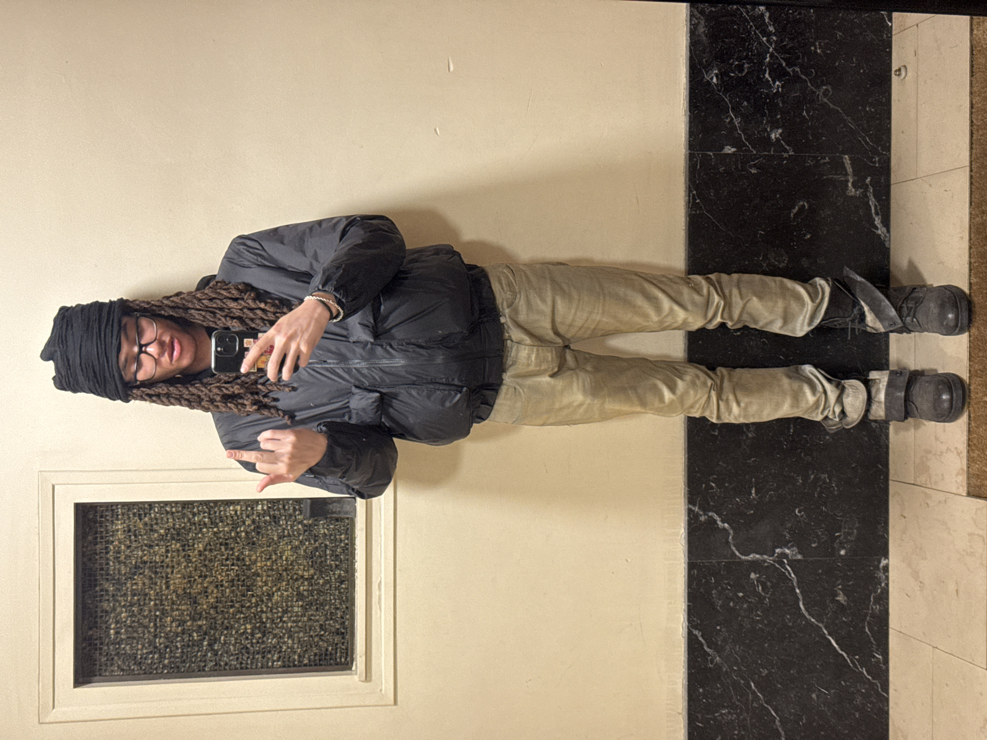 An individual stands centered in an interior hallway or room, situated in Paris, France. The person, with medium-toned skin, wears a black fabric wrapped around their head, partially covering long brown dreadlocks. They have rectangular dark-rimmed glasses and a neutral facial expression with slightly parted lips. The subject is dressed in a black puffer jacket with a visible zipper, light beige or faded brown slim-fit pants, and dark grey or black boots secured with straps. The left boot has a thin, light-colored object tucked into its side. A silver-colored bracelet is worn on the left wrist. The subject holds a dark-colored smartphone in their right hand, angled towards the viewer as if taking a selfie. Their left hand is raised, extending the middle finger.
The background consists of a two-toned wall. The upper section is a plain, light beige color. The lower section features dark, possibly black, marble paneling with prominent white veining, extending approximately from the floor to knee height. In the upper left quadrant of the image, a rectangular window or opening with a light-colored frame is visible. The window is covered by a textured, woven screen, appearing mottled in dark gold and brown tones. The floor at the very bottom is tiled with light-colored squares, transitioning to a darker, possibly brown, carpet or mat directly under the subject's feet.