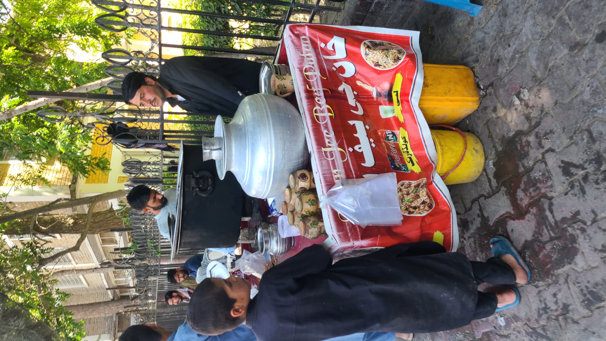 In Quetta City Tehsil, Pakistan, a street food vendor stands by his cart, adorned with a red sign in Urdu, featuring images of food. The cart holds a large silver container, small wooden cups, and plastic bags. Two men are visible: one, dressed in black, stands behind the cart, and another man stands to the left. A boy in black stands in front of the cart. Yellow buckets support the cart. The scene is on a paved street, with buildings and trees in the background, partially obscured by a wrought-iron fence.