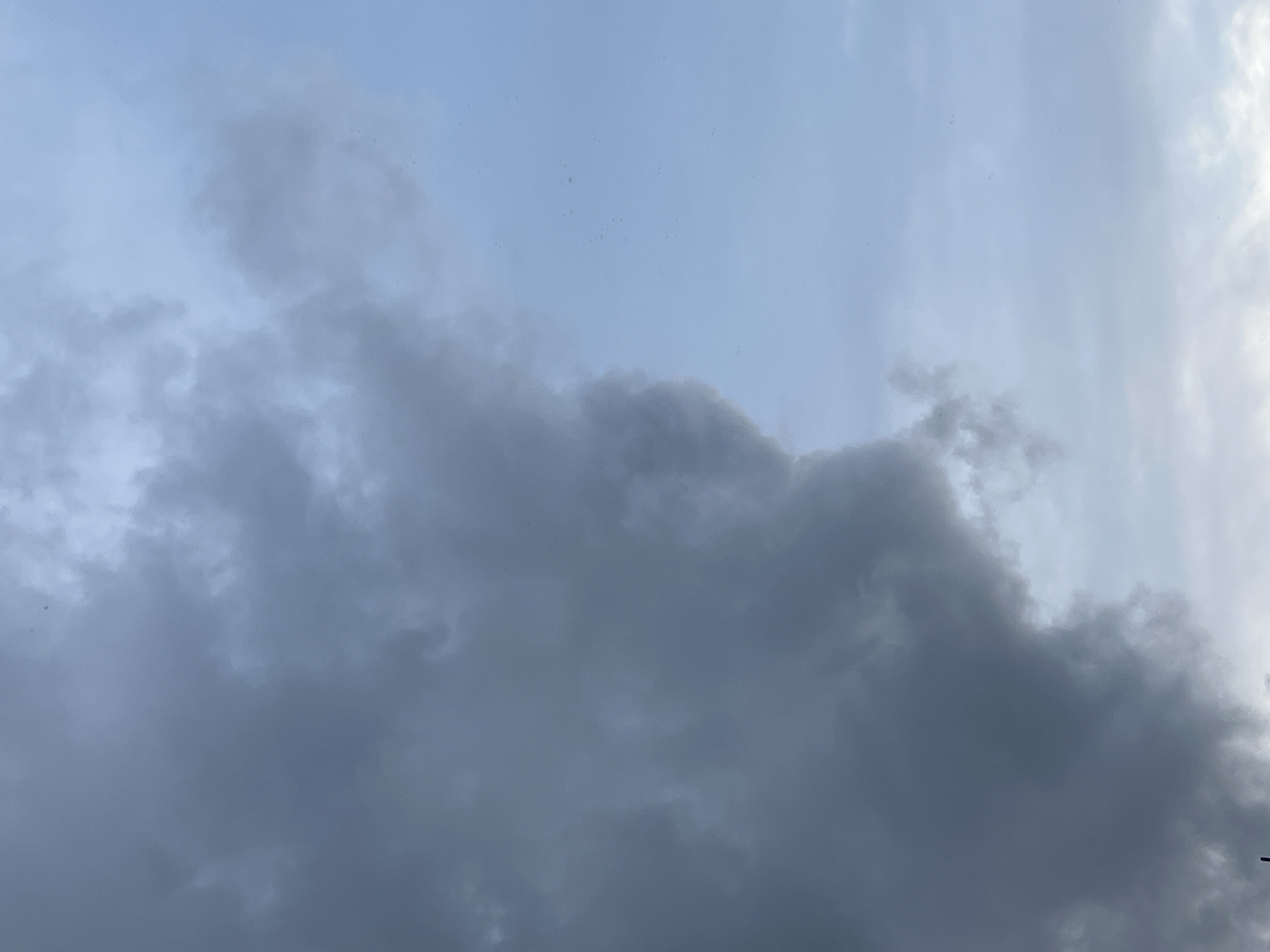 A low-angle, vertical shot captures a portion of the sky. The left side of the frame is dominated by dark, textured storm clouds, their dense mass casting a shadow. To the right, the sky transitions to a lighter, overcast blue, with thin, wispy clouds scattered across it. The overall impression is one of an approaching storm. There are no visible people, structures, or other discernible objects in the frame. The location is the city of Mochishche, Russia.