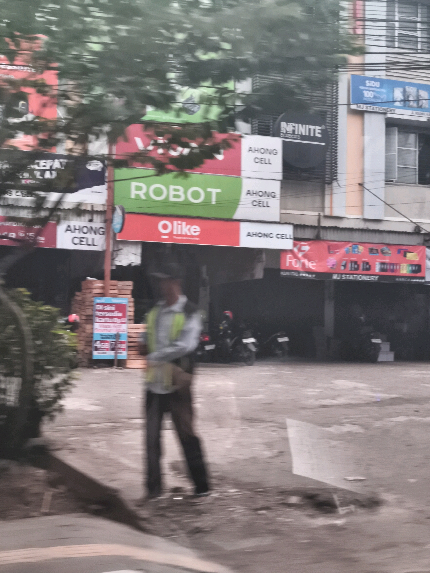 The image depicts a street scene in Palembang, Indonesia, characterized by a blurred motion effect, suggesting the photograph was taken from a moving vehicle. In the foreground, a person in a safety vest and dark pants stands near a stack of wooden pallets. The background features several shops and businesses, indicated by signage. Visible signs include "ROBOT," "Olike," "AHONG CELL," "INFINITE," and "SIDU." The buildings have multiple stories and some visible windows. Motorcycles are parked in front of some of the establishments. The ground appears to be paved, with some areas of dirt and puddles, possibly indicating recent rain. Trees with dense foliage are present on the left side of the image, partially obscuring the view of the buildings.