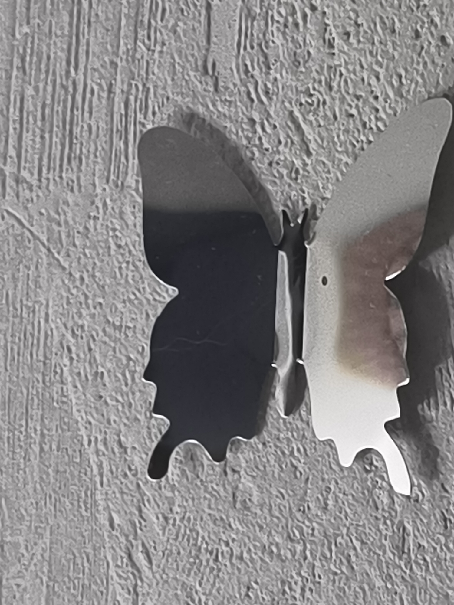 The image shows a close-up of a metallic butterfly decoration affixed to a textured, grey wall. The butterfly is a stylized, three-dimensional object with its wings extended. The left wing is primarily dark, appearing black or a very deep grey, reflecting some of the textured wall. The right wing is a lighter, reflective metallic surface with subtle reddish-brown and white marbling. A small dark dot, possibly a rivet or a design element, is visible on the right wing. The wall behind the butterfly has a rough, irregular texture with vertical striations. The lighting casts a distinct shadow of the butterfly onto the wall, accentuating its form. There are no people or other objects visible in the frame. The scene is indoors, likely in a residential or commercial space, given the wall decoration. No specific location in Klaipėda, Lithuania can be determined from the visual information provided in the image.