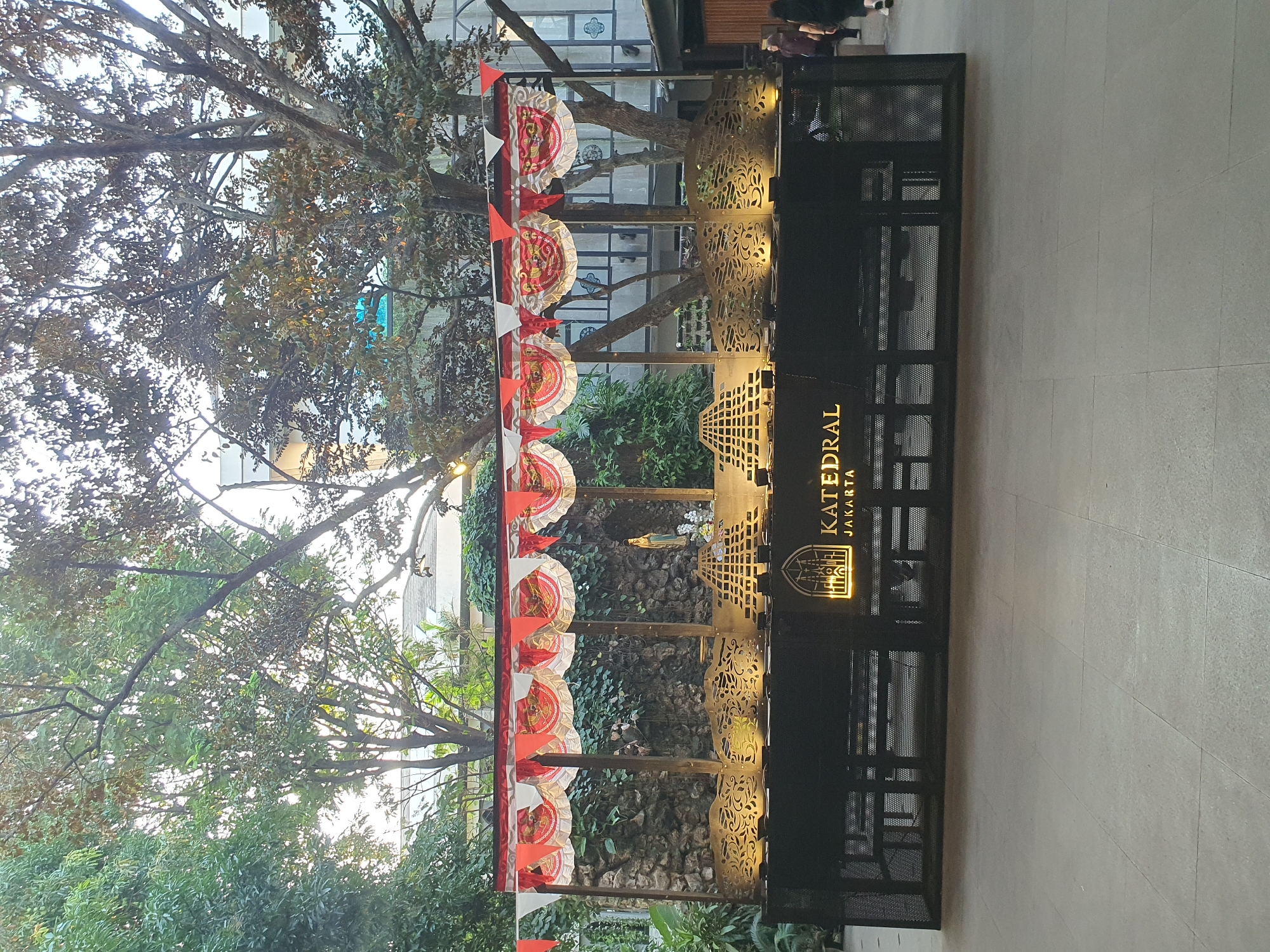 In an outdoor courtyard setting in Central Jakarta, Indonesia, a temporary display is set up. The display features a dark counter with a backlit logo reading "KATEDRAL JAKARTA" and an image of a cathedral. Above the counter, a structure is adorned with red and white pennant banners and decorative circular ornaments. Behind the display is a textured stone wall covered in ivy, and to the right, a portion of a building with large windows is visible. Several trees with dense foliage are present in the background, and a few people are walking in the far right of the frame. The ground is paved with large, square tiles.
