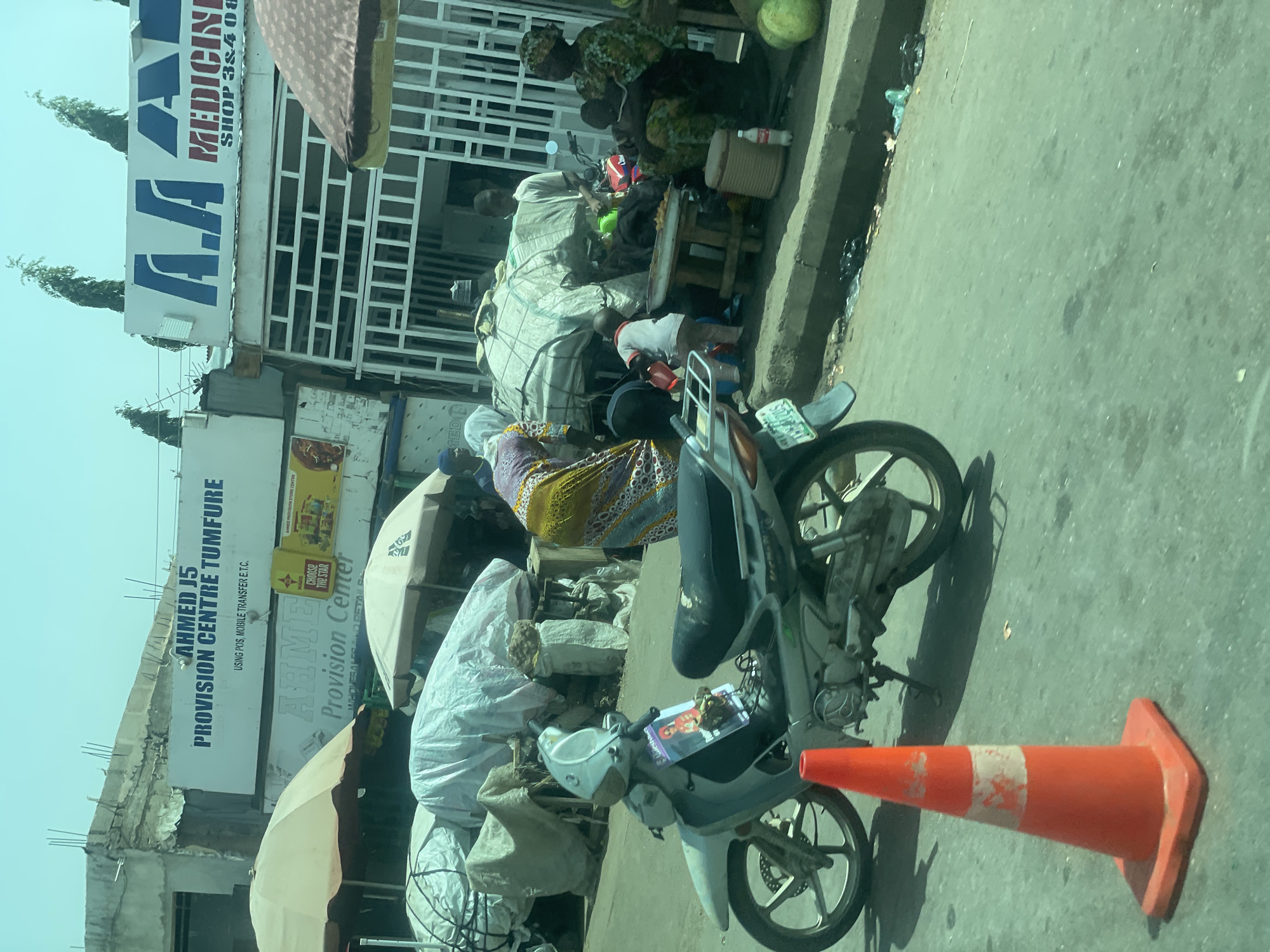 This image, taken in Gombe, Nigeria, depicts a bustling street scene. Several people are visible, some appearing to be vendors or customers. In the foreground, a motorcycle is parked, with an orange traffic cone lying on the ground next to it. The setting appears to be a market or commercial area, with several storefronts and stalls.
One prominent sign reads "AHMED J5 PROVISION CENTRE TUMFURE USING POS, MOBILE TRANSFER E.T.C." and another nearby sign displays "AHME Provision Center" with "CHOOSE THE STAR" below it. There is also a sign that says "A.A MEDICIN SHOP 38408". These signs suggest a commercial hub with various goods and services available.
The people are dressed in typical attire, and their interactions are not clearly discernible due to the image's resolution and angle. The scene is lit by natural daylight, indicating it was taken during the day. The presence of the motorcycle and the various goods for sale suggest ongoing economic activity. The overall impression is one of a vibrant, everyday scene in a Nigerian marketplace.