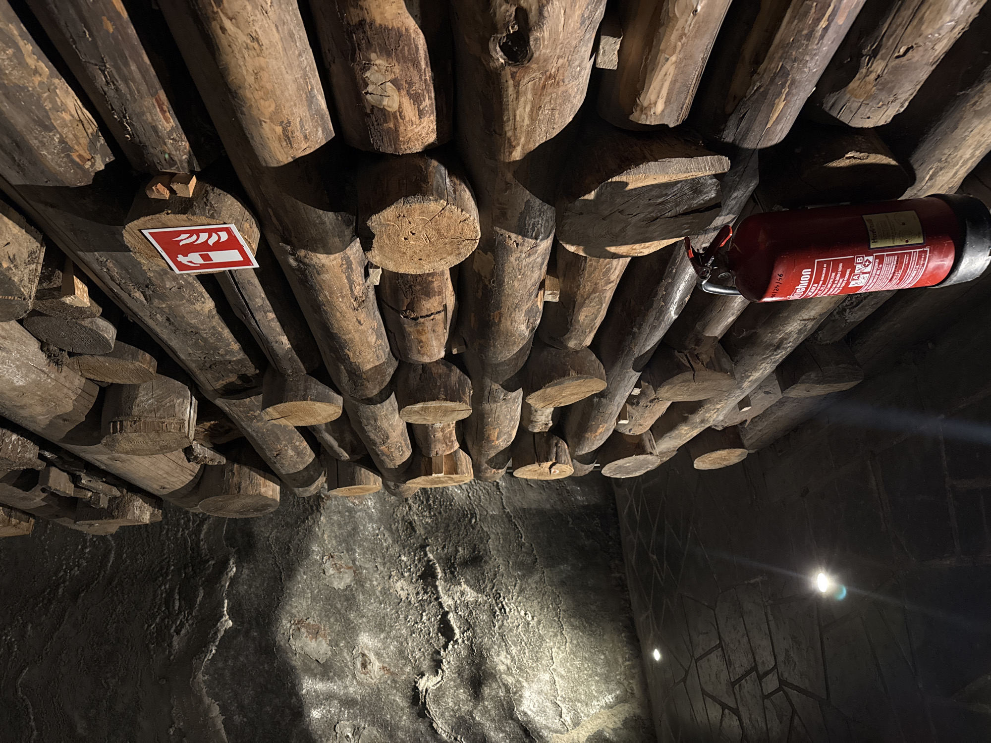 The image depicts an underground space in Wieliczka, Poland. The ceiling is constructed of round wooden logs, supported by upright wooden posts. A red fire extinguisher is mounted to the logs on the right side. A red and white fire safety sign is also visible on the logs. The walls are rough and made of grey stone. A light source illuminates the scene from the lower right, casting shadows. No people are visible.