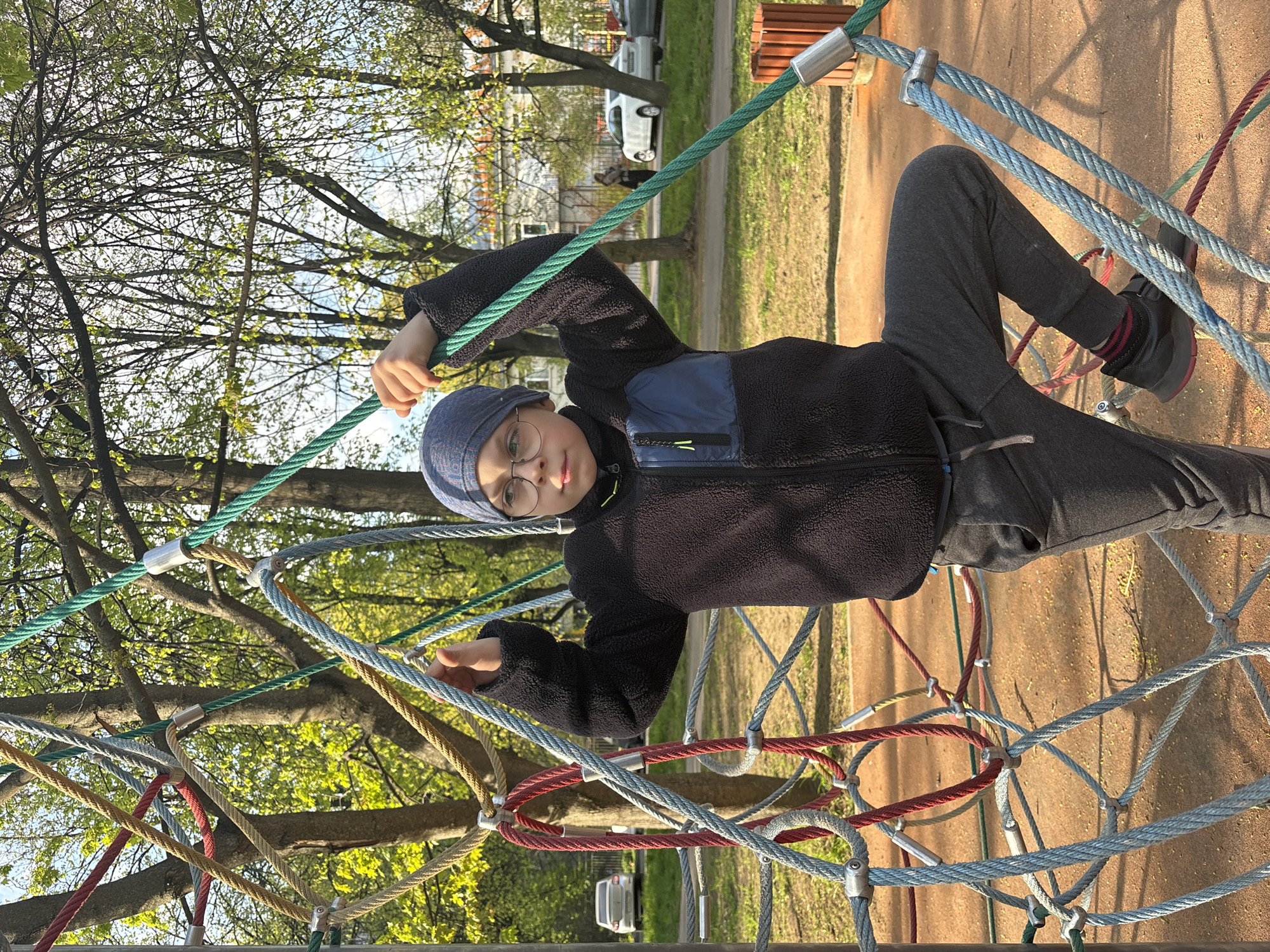 A child is climbing on a rope playground structure in what appears to be a park. The child is wearing glasses, a dark-colored fleece jacket, a hat, and dark pants. The playground structure consists of ropes of various colors (green, blue, red, brown) interconnected to form a climbing net. Trees with green leaves are visible in the background, suggesting springtime. Buildings and parked cars can be seen in the distance. The location is Saint Petersburg, Russia.