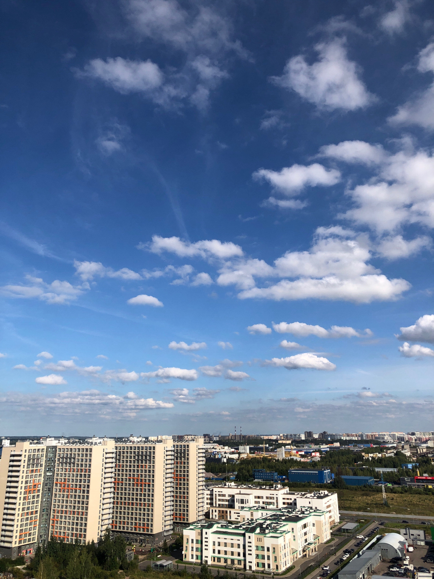 This is a vertical, high-angle shot of a modern residential area in Lavriki, Russia, under a bright blue sky dotted with scattered white cumulus clouds. The foreground features several tall, cream-colored apartment buildings with numerous windows, some accented with orange vertical stripes. To the right, a complex of lower, white buildings with green accents is visible, bordering a road with parked cars. Beyond the residential buildings, a dense green forest separates them from an industrial zone with various factory-like structures, including large blue buildings. In the far distance, a sprawling cityscape with numerous buildings of varying heights stretches across the horizon. The overall scene depicts a developing urban landscape with a mix of residential, industrial, and natural elements. There are no people or animals visible in the image.