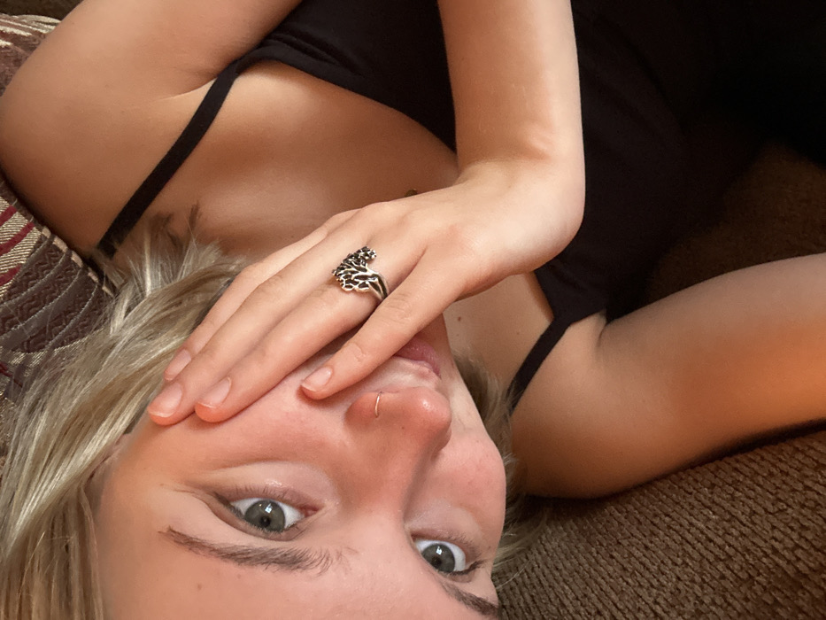 A woman reclines on patterned fabric, possibly a cushion or couch. She wears a black tank top and has fair skin and blonde hair. Her right hand, adorned with a silver ring in the shape of a lion, partially obscures her face. Her eyes are wide and green. A small silver hoop adorns her left nostril. The image is taken from a low angle, creating an intimate and somewhat disorienting perspective.
