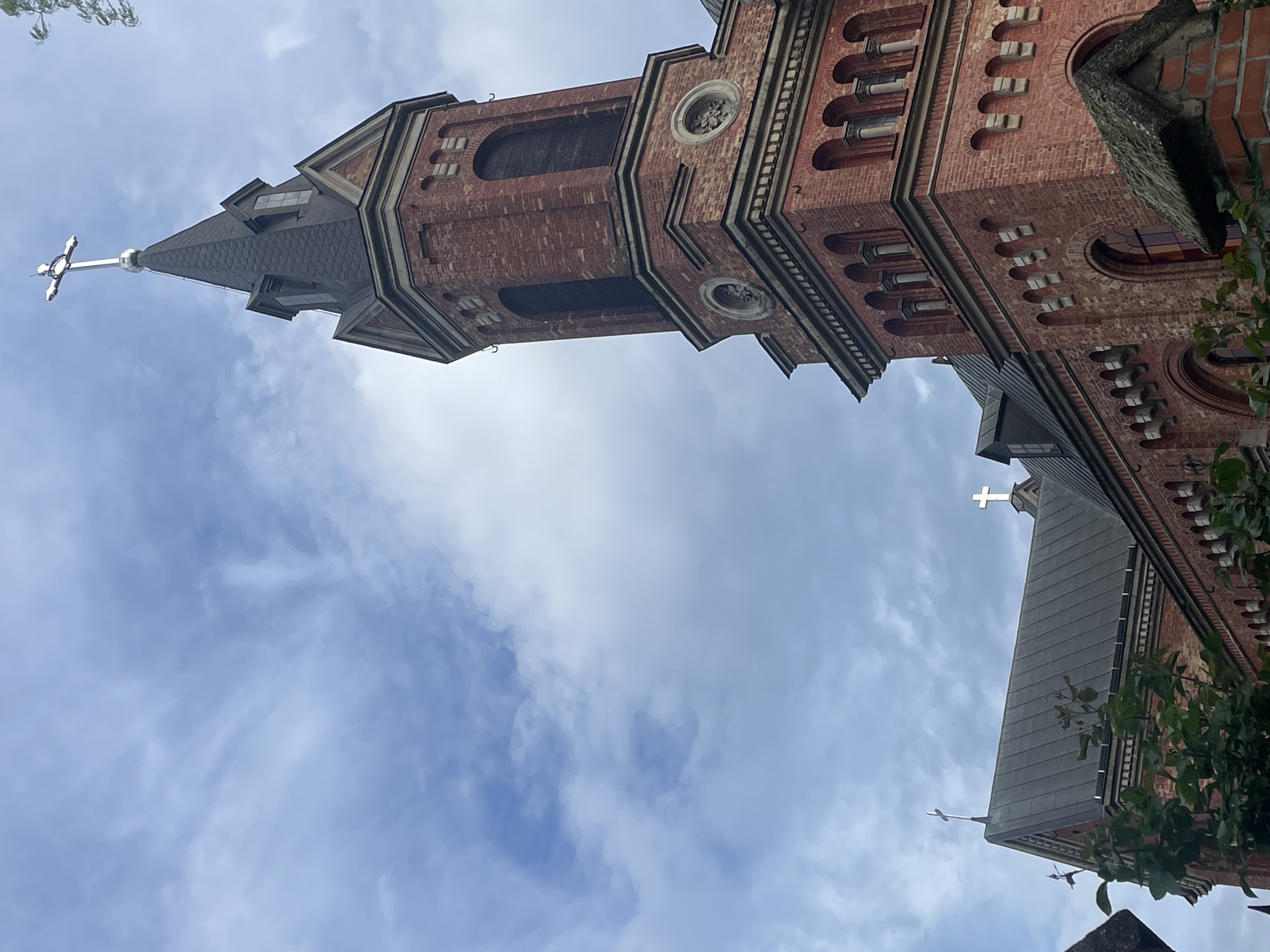 The image shows a tall, red brick church tower against a cloudy sky. The tower has a pointed, dark roof with a cross at the very top. There are architectural details such as arched windows and circular embellishments. Part of another section of the church is visible on the right, also featuring a cross. Some greenery is visible in the lower right corner. The location is inferred as Mykolaiv, Ukraine, based on the original prompt.
