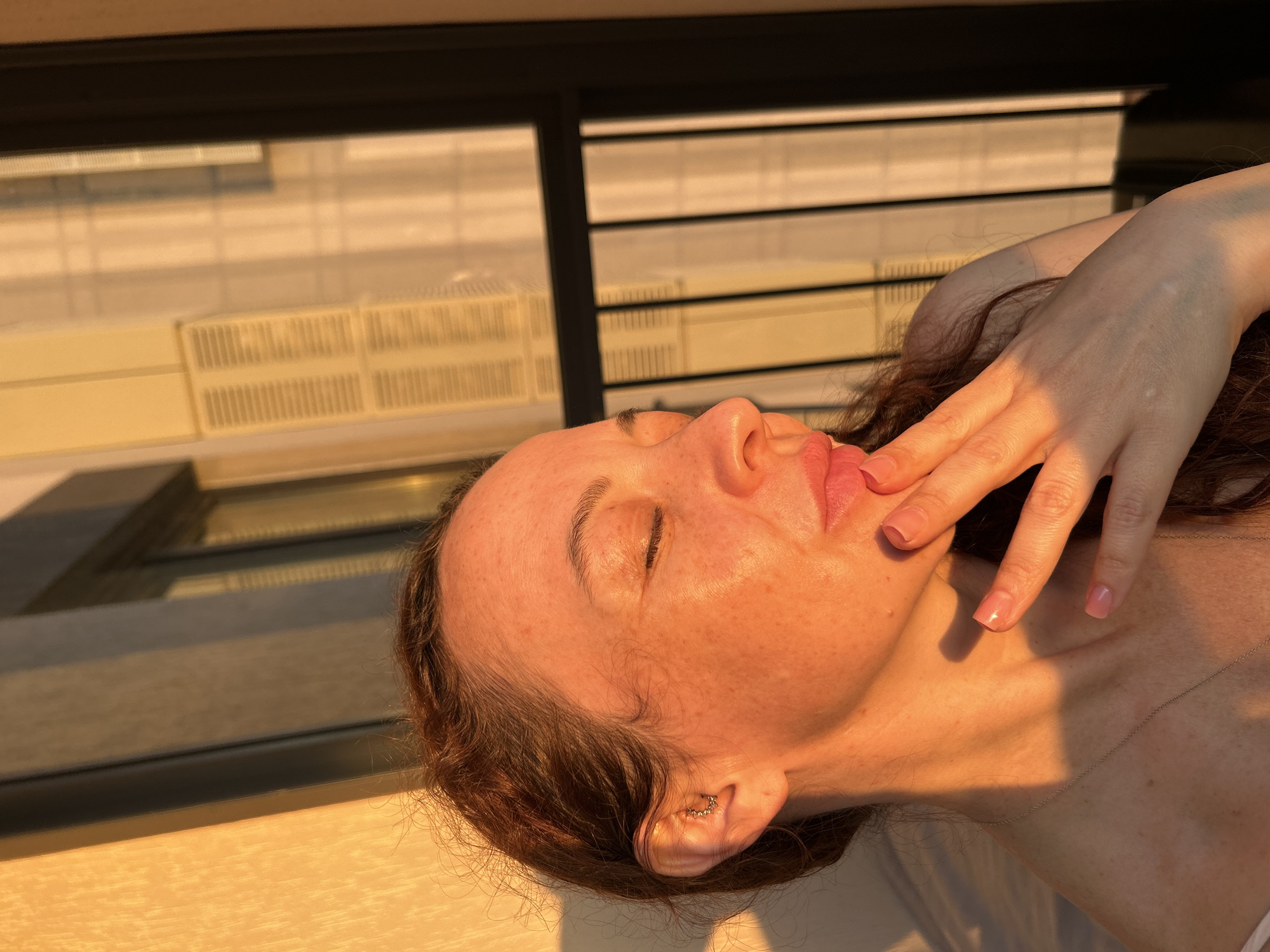 A medium close-up shot captures a woman in profile with her eyes closed, bathed in the warm glow of sunset. Her head is tilted slightly, and her right hand is raised, with her fingers gently touching her lips. She has long, auburn hair that cascades over her shoulder. The background features a building with vertical window panes and louvers, suggestive of an urban setting. The lighting indicates the golden hour, casting long shadows and highlighting the textures of her skin and the architectural elements. There is no discernible information about the location being Yekaterinburg, Russia.