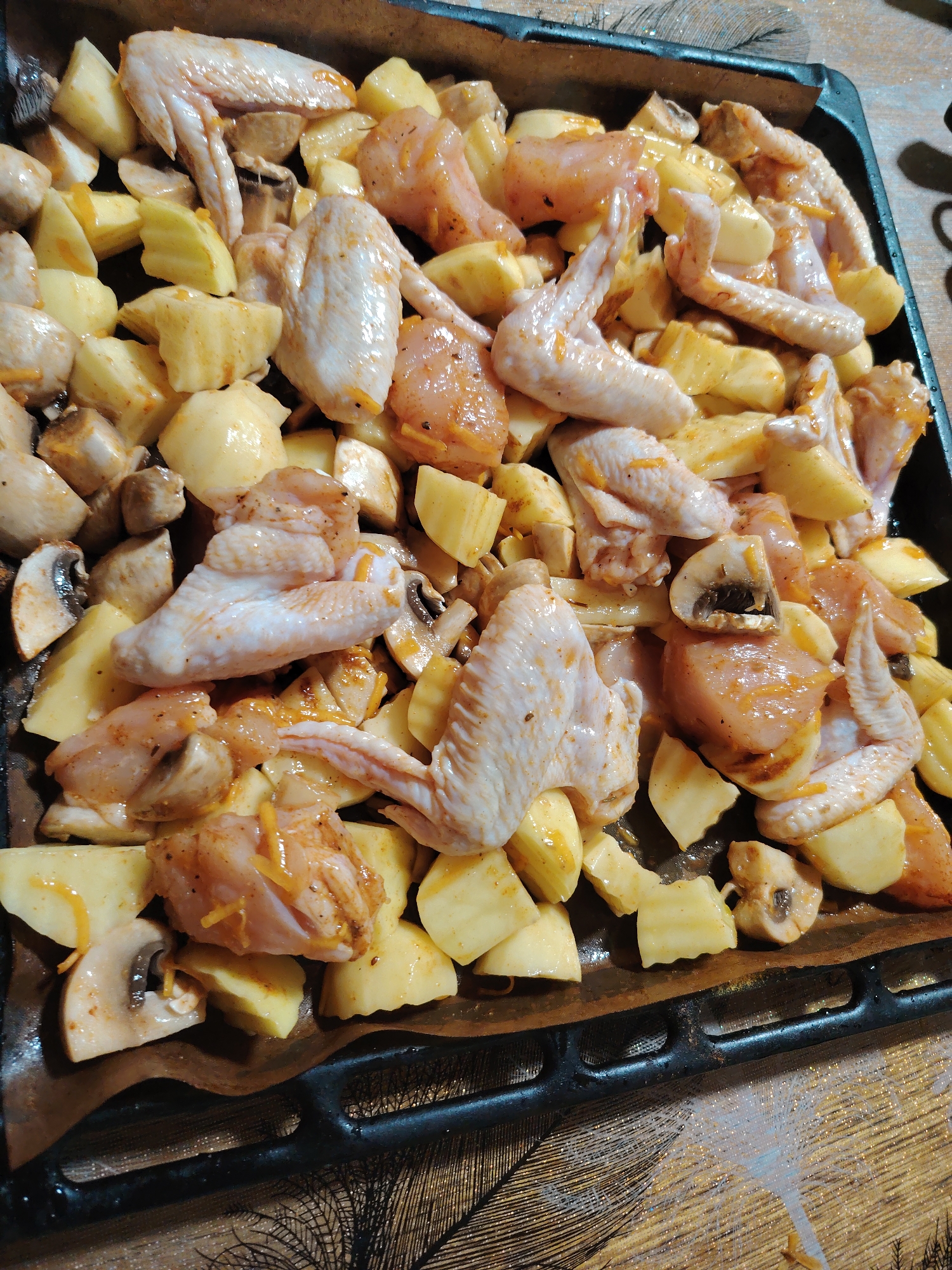 This is a close-up, overhead shot of a baking tray filled with raw chicken wings and chopped potatoes, interspersed with sliced mushrooms. The chicken wings appear to be seasoned with a reddish-orange spice mix. The potatoes are cut into roughly uniform, bite-sized pieces. The mushrooms are sliced, with some showing visible gills. The entire mixture is arranged on a dark baking tray, possibly lined with parchment paper, which is placed on a patterned tablecloth. The lighting suggests an indoor setting, likely a kitchen, with no specific cues for time of day or weather. The scene depicts food preparation, specifically ingredients for a meal ready to be baked or roasted. There is no visible text in the image. The location context of Rostov-on-Don, Russia, does not provide any visually identifiable details within the image itself.